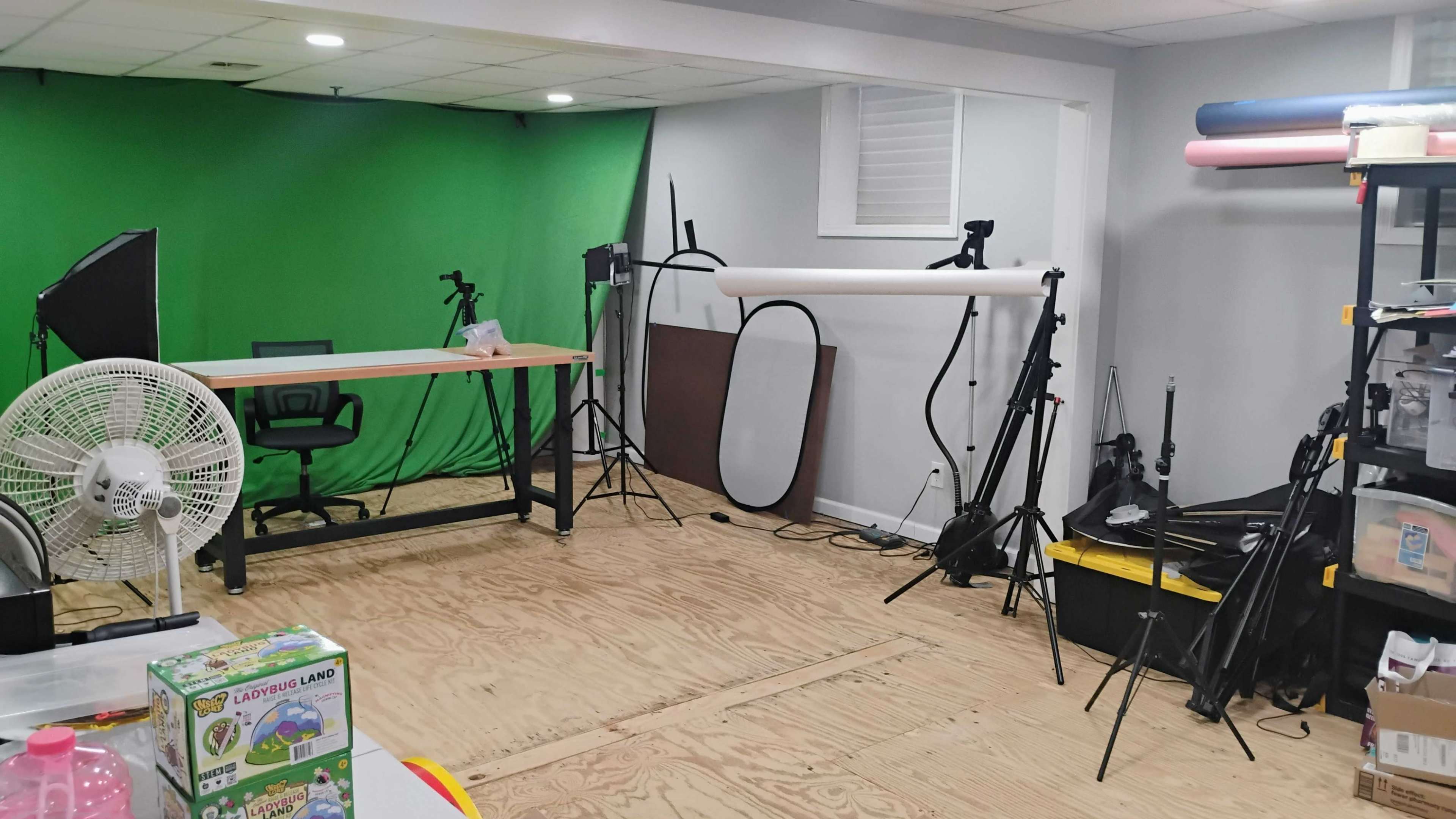 The image shows a studio space with a green screen, a desk and chair, various lighting equipment, and shelves with items in the foreground.