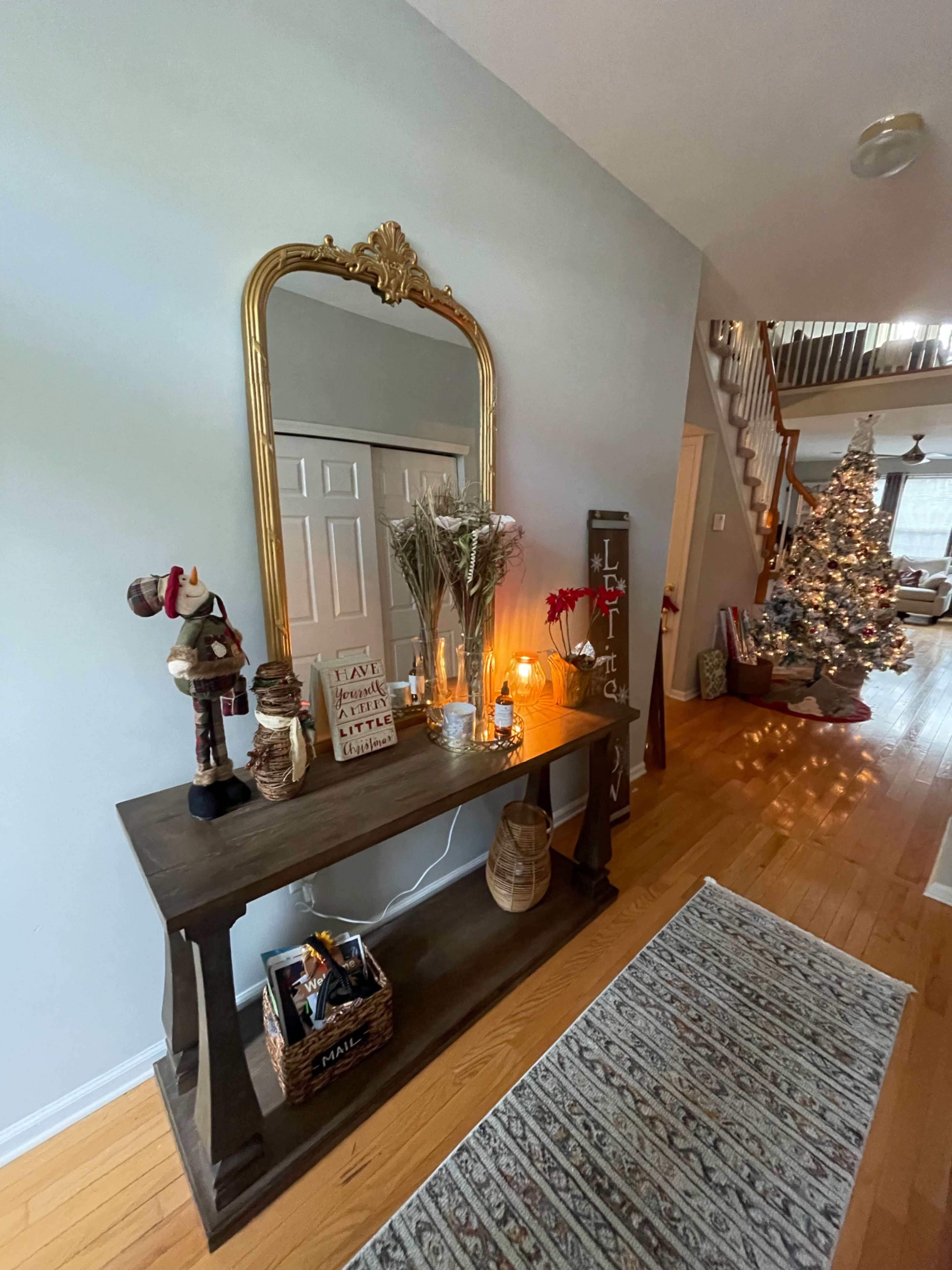 A console table adorned with decorative items, a large mirror, and candles is positioned against a light-colored wall in a hallway, with a Christmas tree visible in the background.