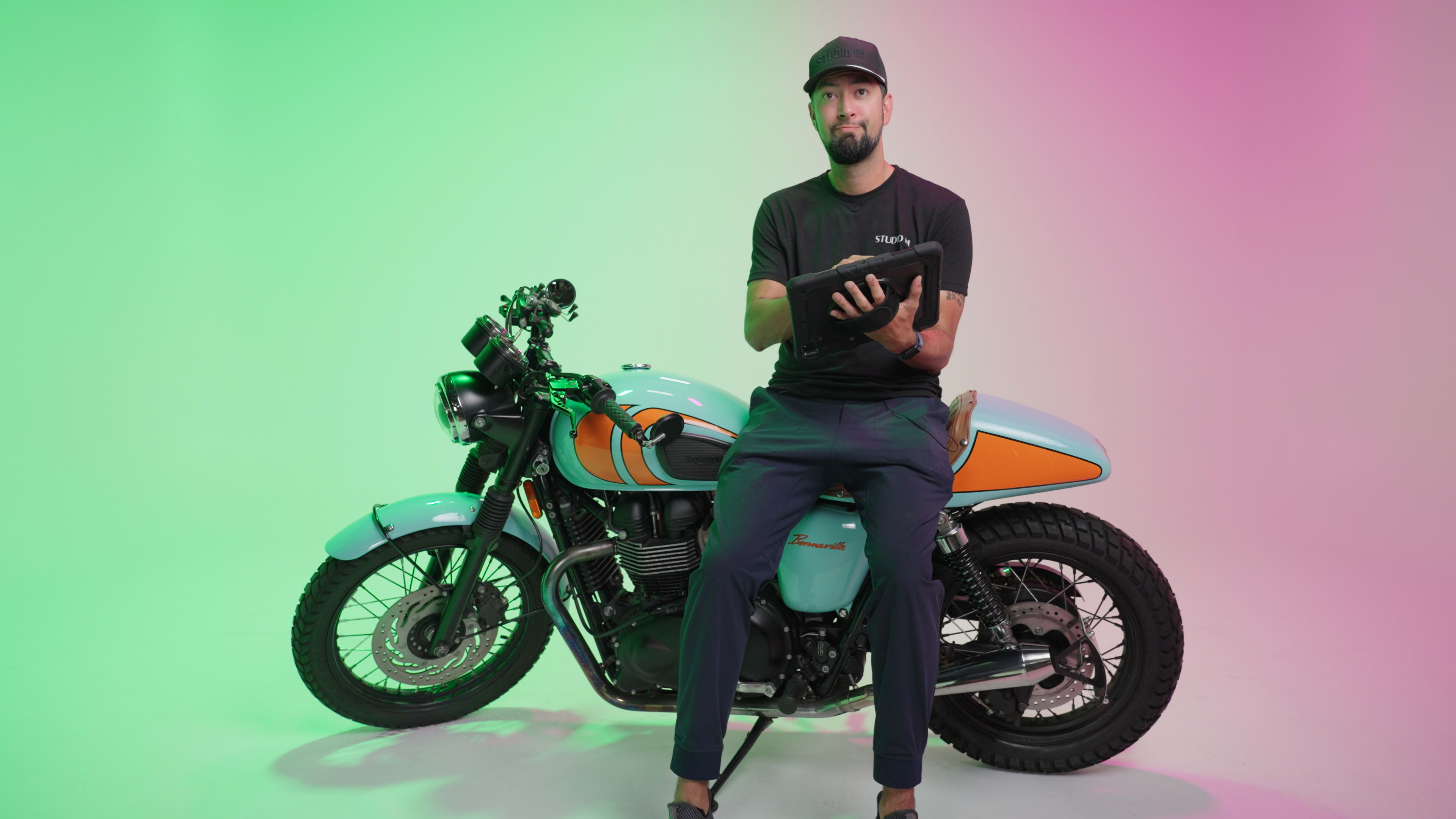 A man sits on a vintage motorcycle against a gradient pink and green backdrop, holding a tablet.