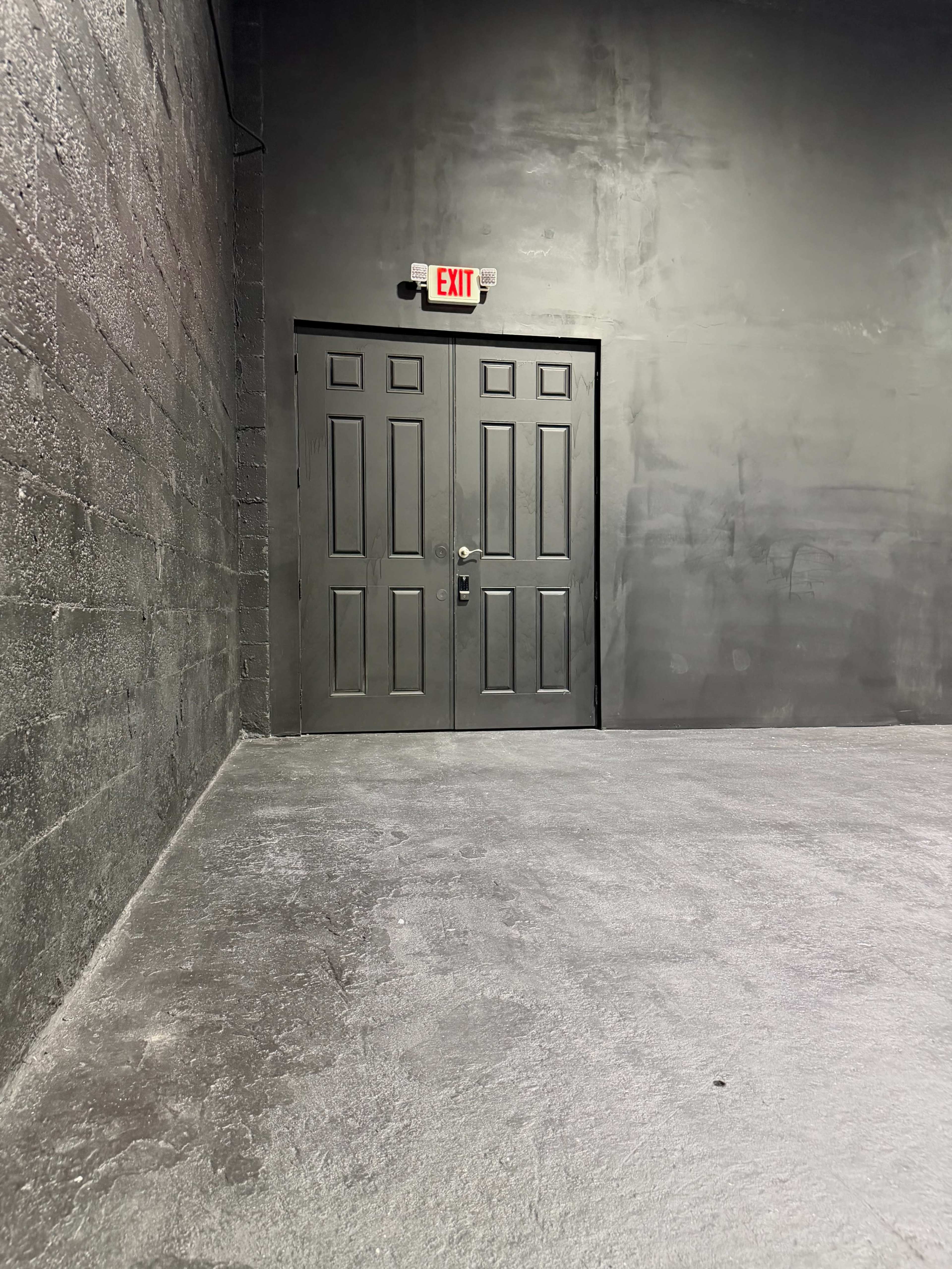 The image shows a pair of gray double doors at the end of a plain, dimly lit room with a concrete floor and gray walls.