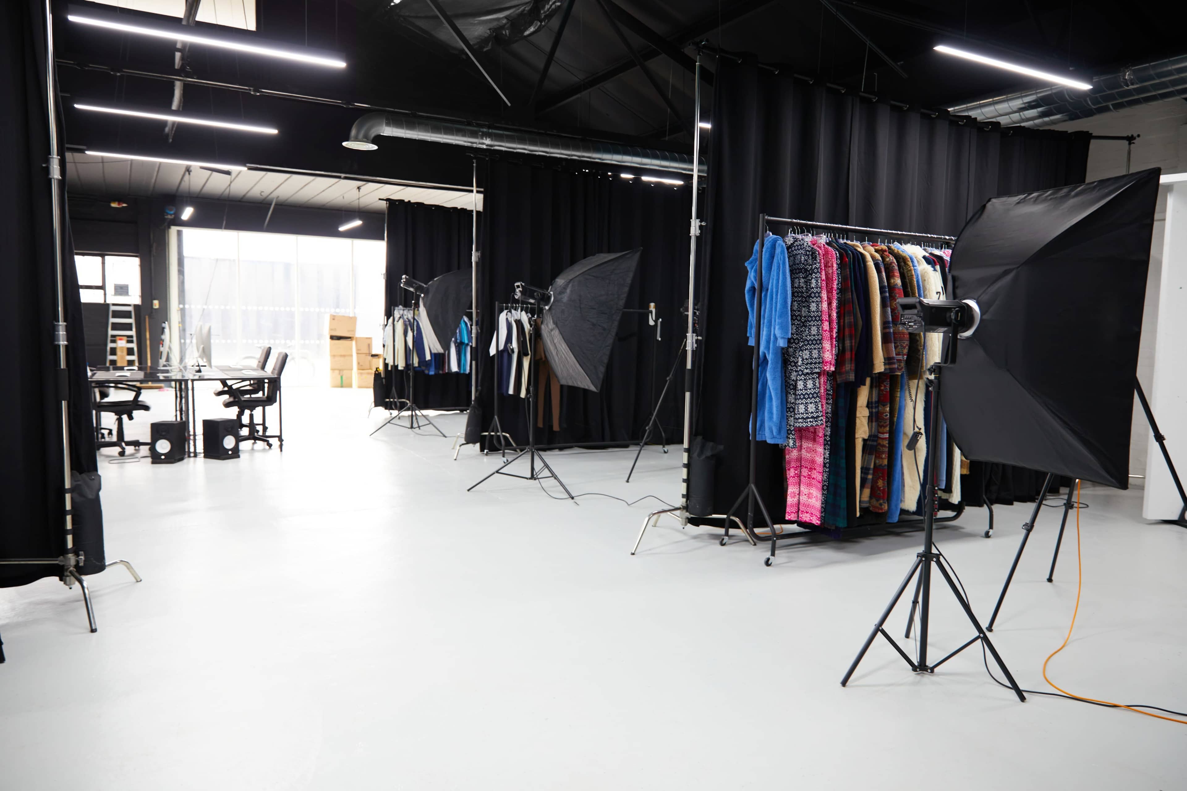The image shows a photography studio with black backdrops, lighting equipment, and clothing racks arranged along the sides.