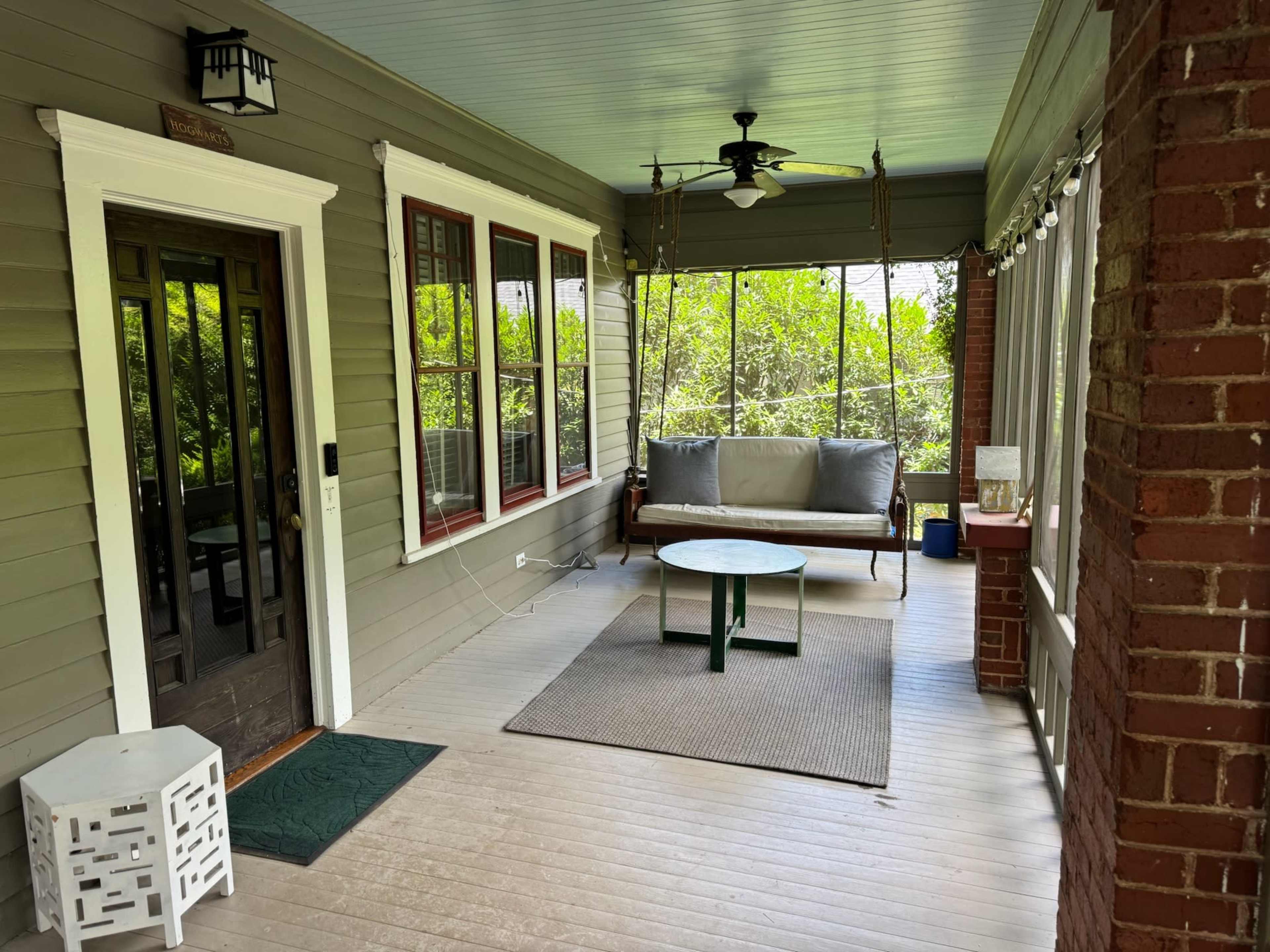 The image shows a porch with a swing seat, a round coffee table, and several windows overlooking greenery.