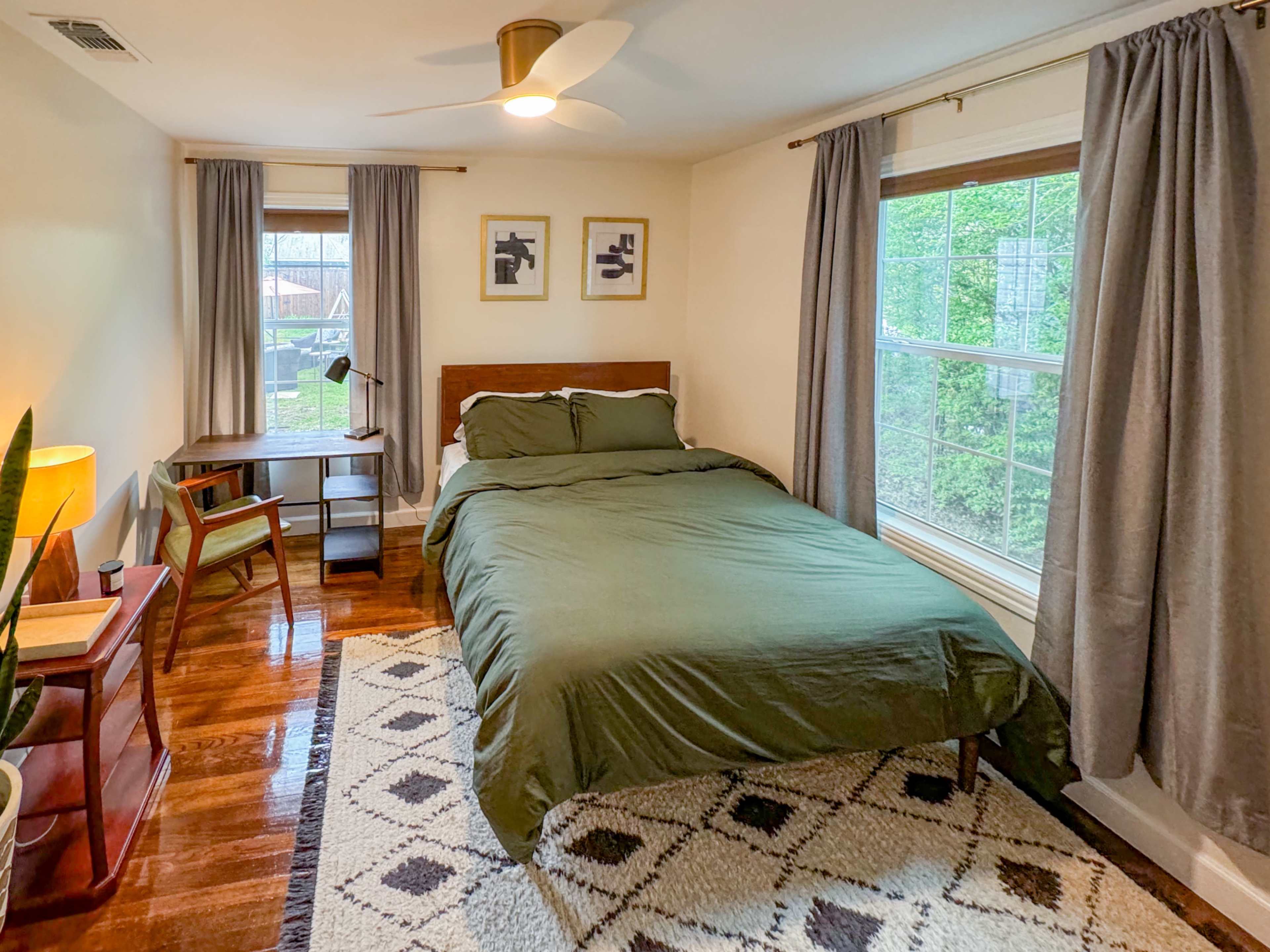The image shows a bedroom with a double bed, a desk and chair, two windows with light gray curtains, and a patterned rug on hardwood floors.