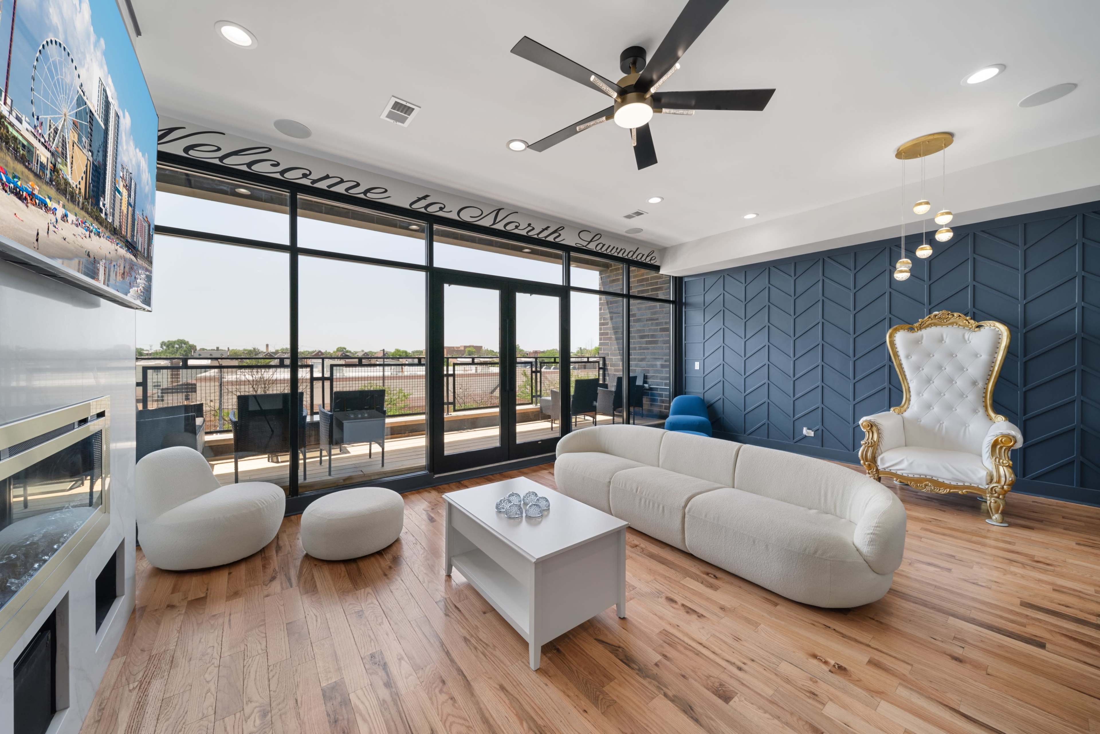 The image shows a modern living room featuring a white curved sofa, a small white coffee table, and a large decorative chair, with large windows offering a view of an outdoor balcony.
