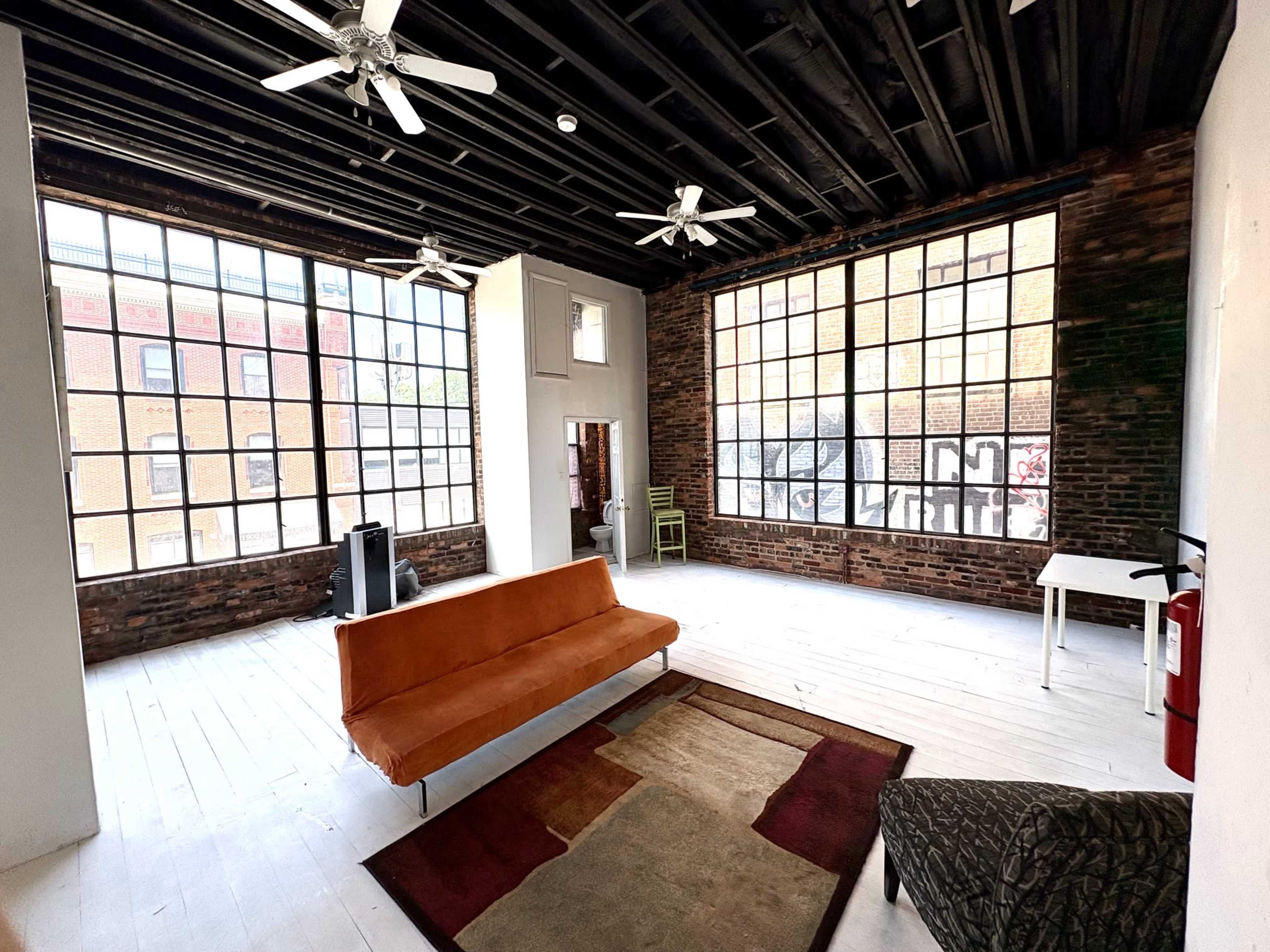 The image shows a spacious room with large windows, exposed brick walls, and minimal furnishings, including a vintage orange sofa and a small white table.
