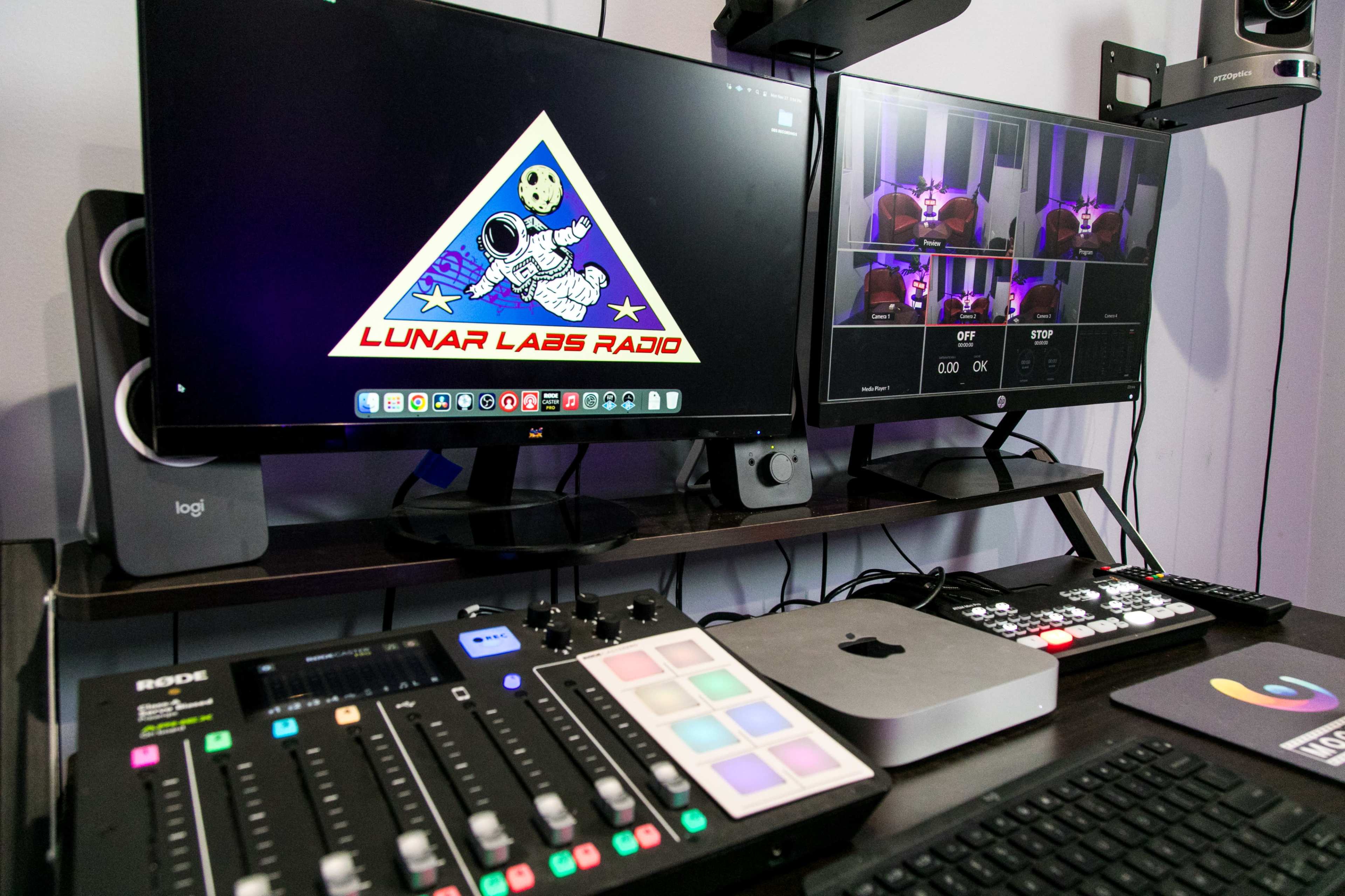 A radio studio setup with two monitors, an audio mixing console, and various control devices on a desk prominently featuring the "Lunar Labs Radio" logo.