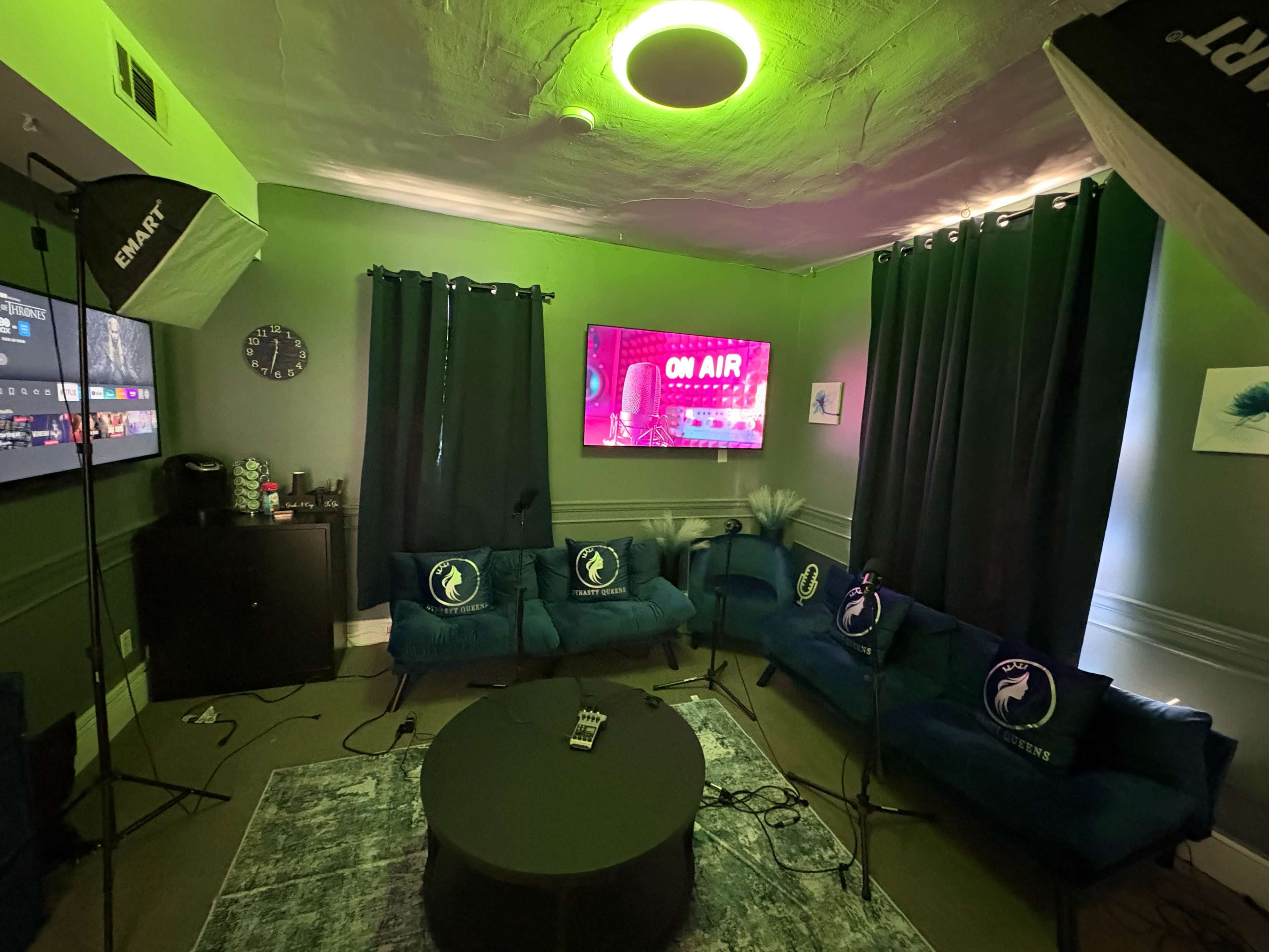 The image shows a cozy studio setup with green walls, two blue couches, a circular coffee table, and lighting that casts a green glow, featuring a wall-mounted screen displaying "ON AIR."