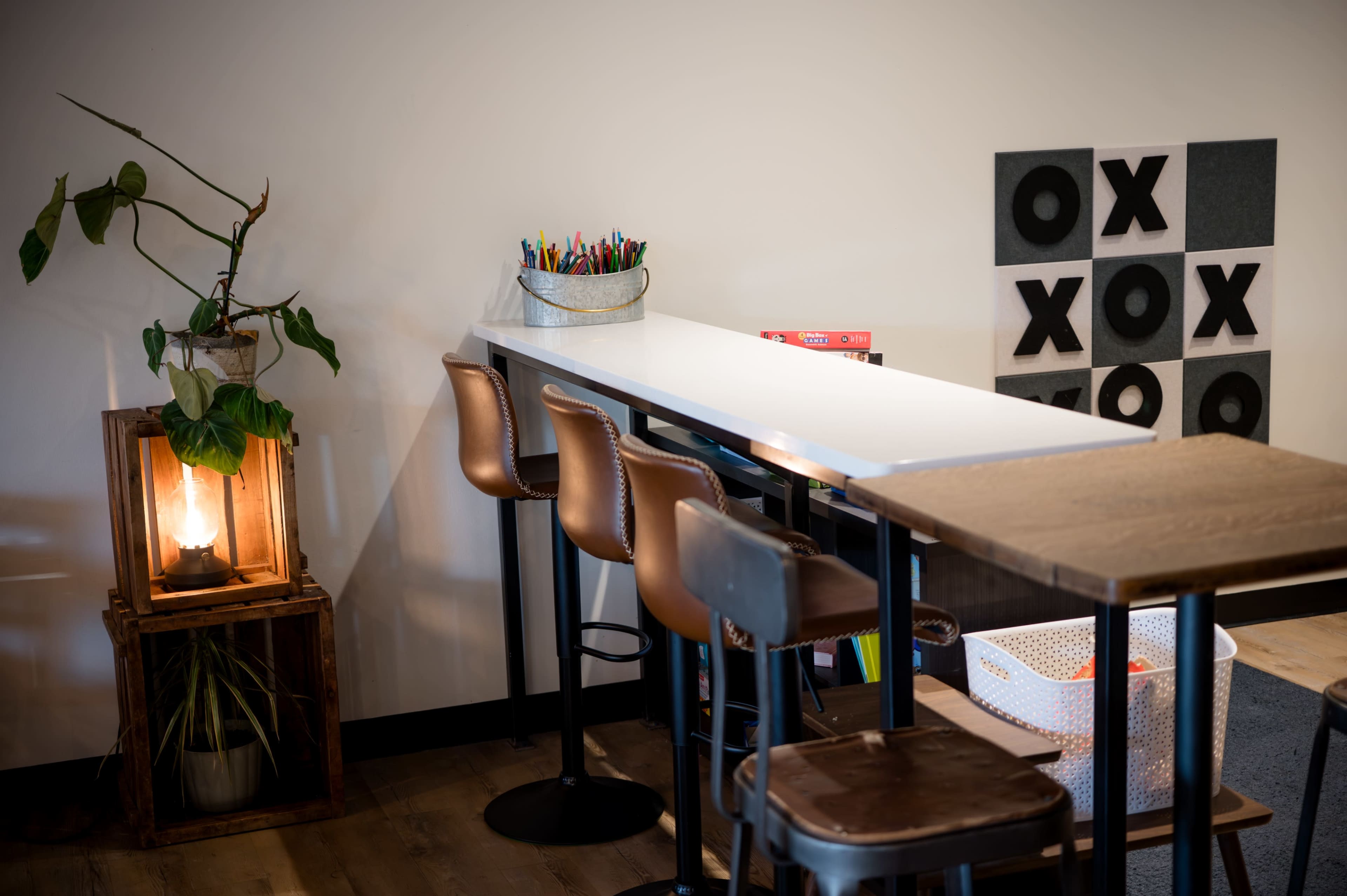 The image shows a modern workspace with a long white counter, brown bar stools, a plant in a wooden box, and a wall featuring a grid of black and white "XOXO" art.