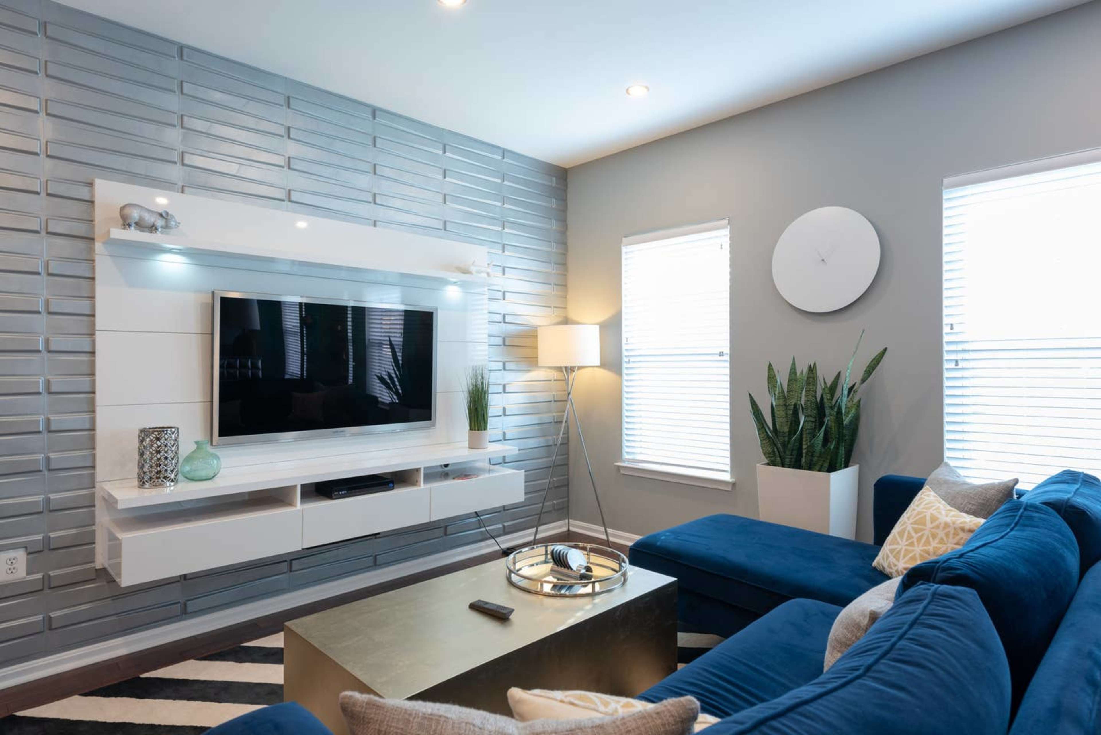 A modern living room with a blue sectional sofa, a wall-mounted television, and large windows with blinds.