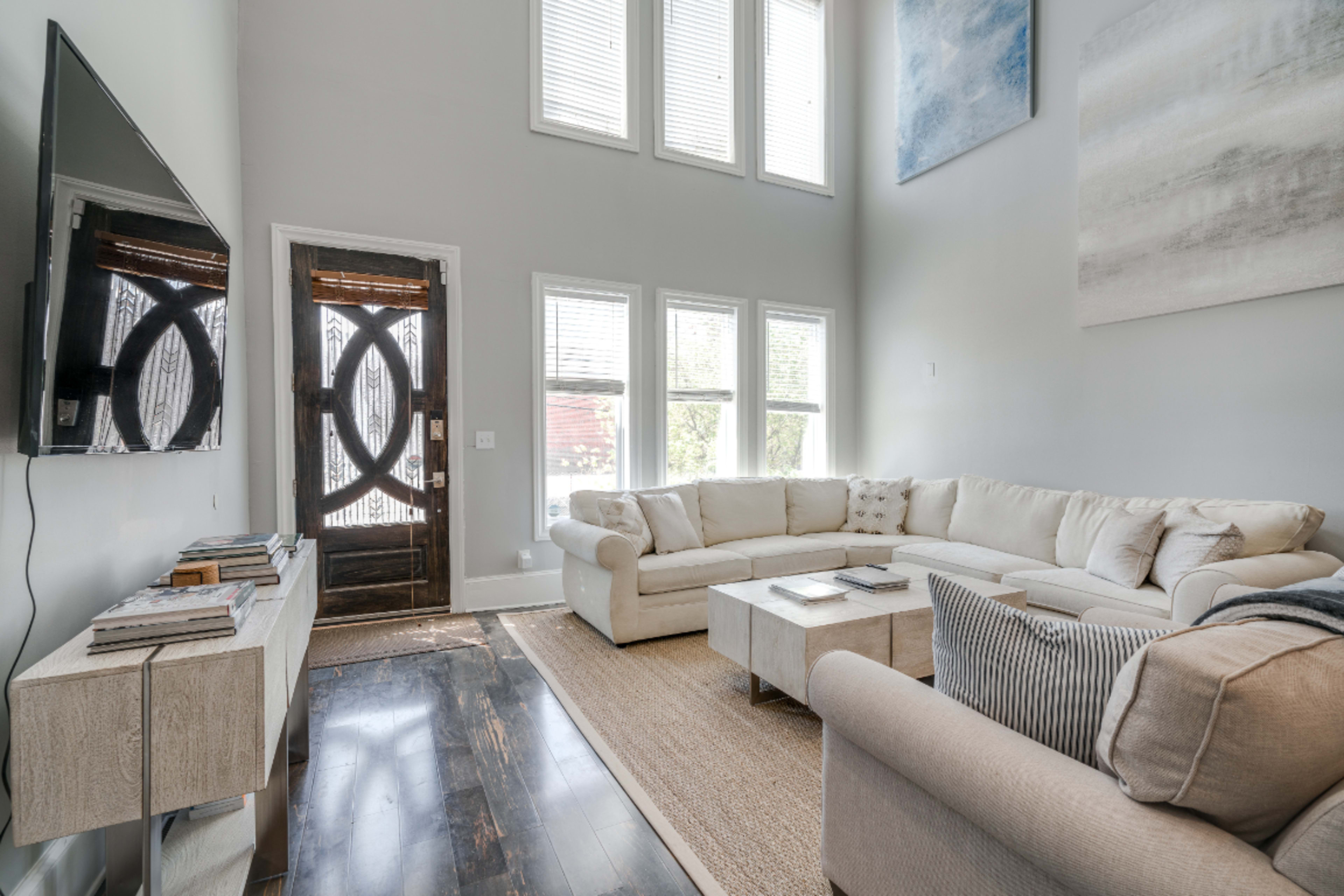 The image shows a modern living room with a large sectional couch, a coffee table, a flat-screen TV mounted on the wall, and bright windows allowing natural light.