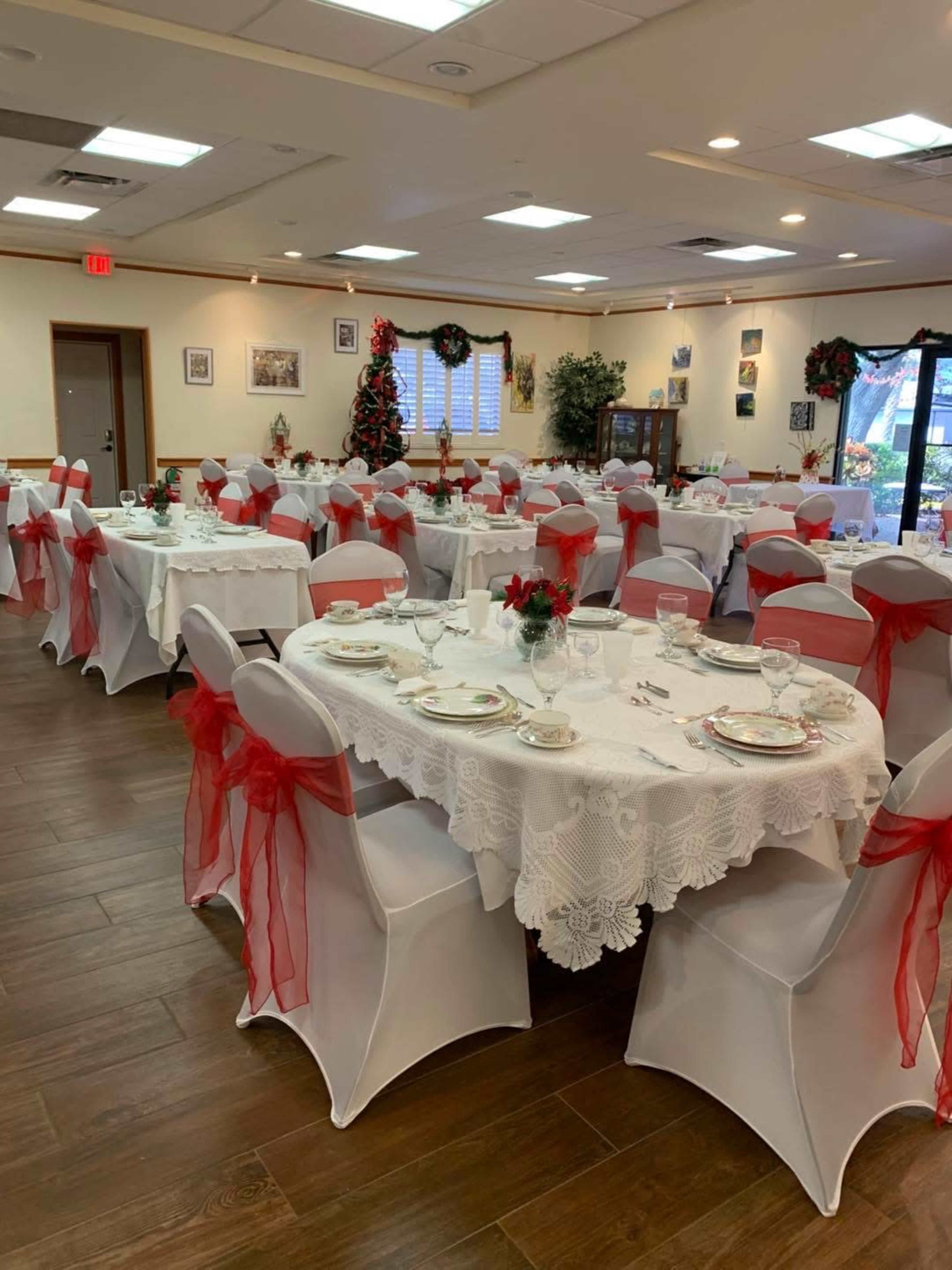 The image shows a banquet hall decorated for a festive event, with tables covered in white lace tablecloths and adorned with red ribbons, along with floral centerpieces.