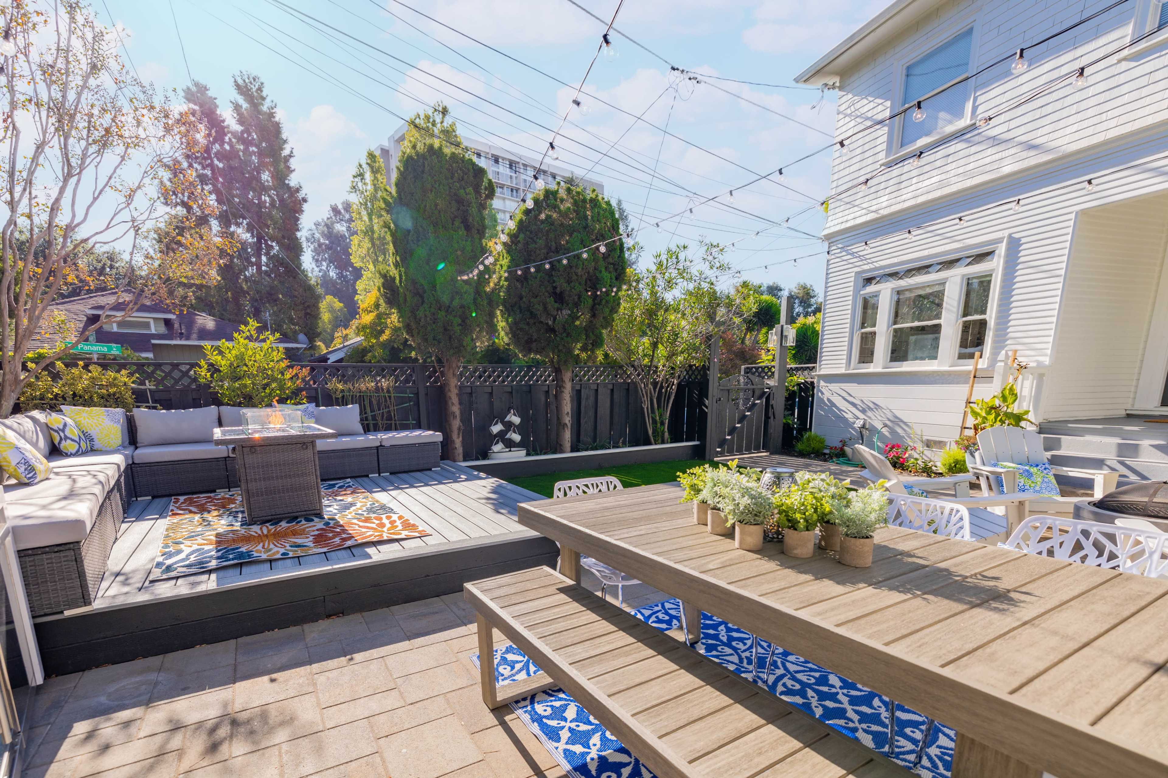 A well-designed backyard patio area featuring a seating arrangement with a fire pit, a dining table with potted plants, and string lights overhead, surrounded by greenery.