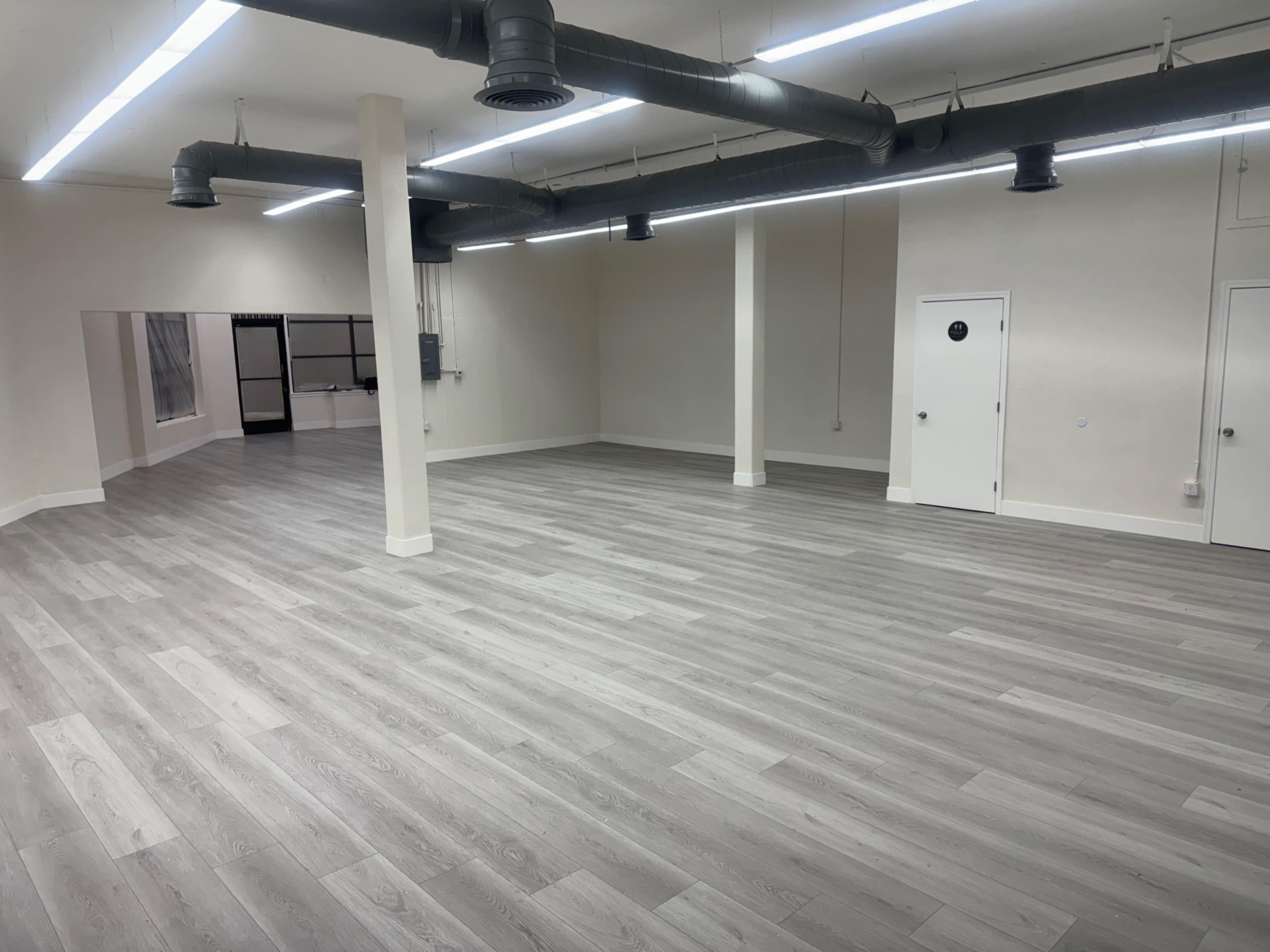 The image shows a spacious, empty room with gray wooden flooring and exposed ductwork on the ceiling.