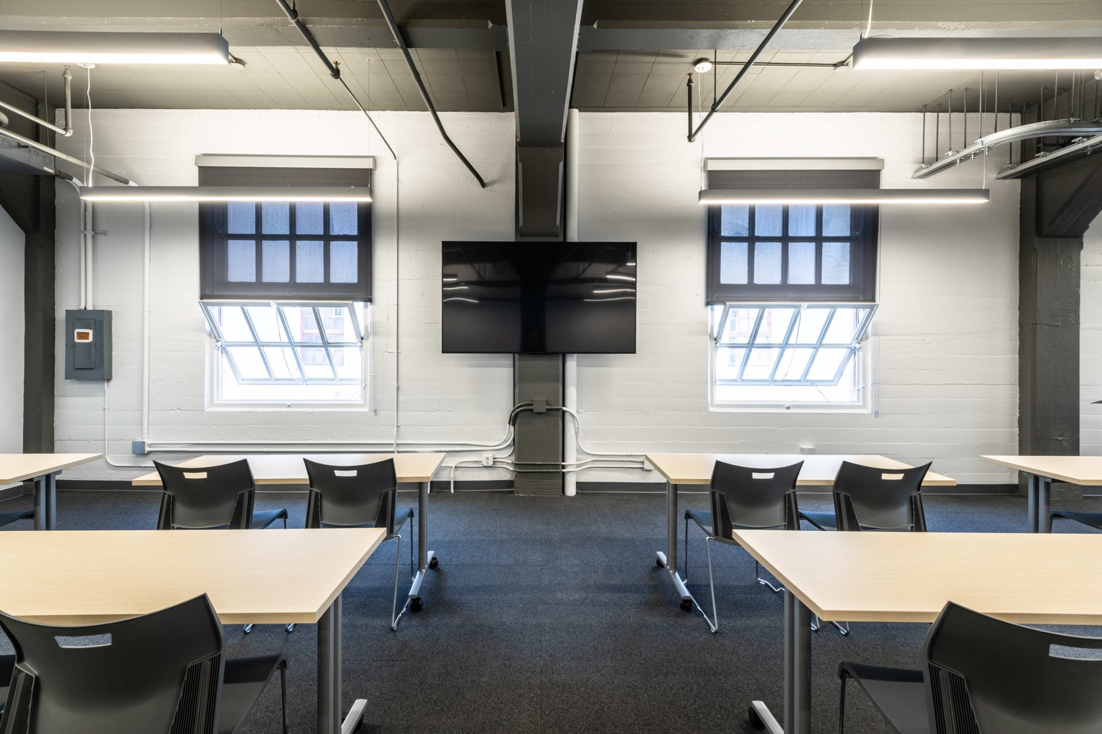 A modern classroom features rows of tables and chairs facing a wall-mounted television, with large windows on either side.