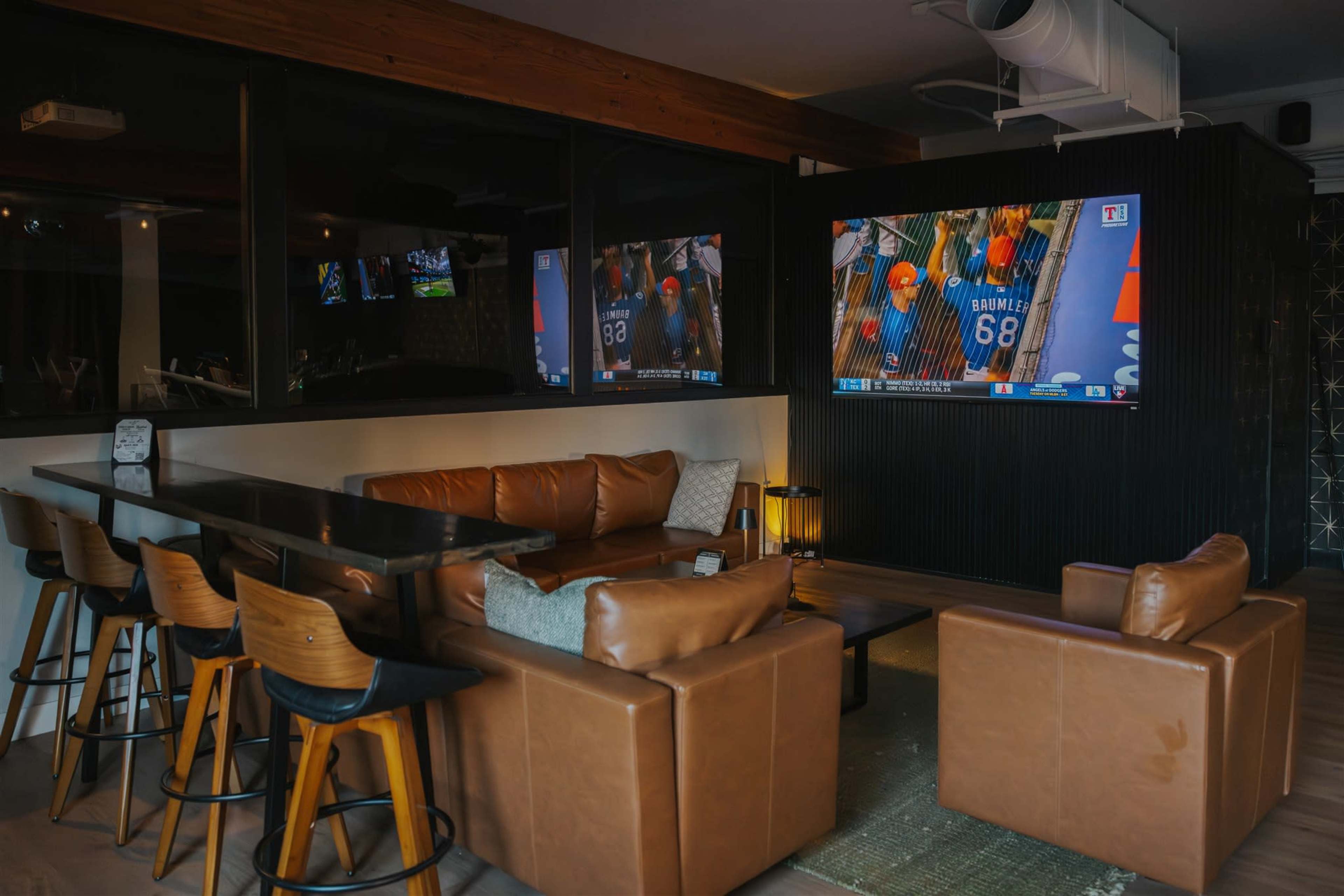 The image shows a modern lounge area with leather sofas, wooden bar stools, and two large screens displaying a sports game.