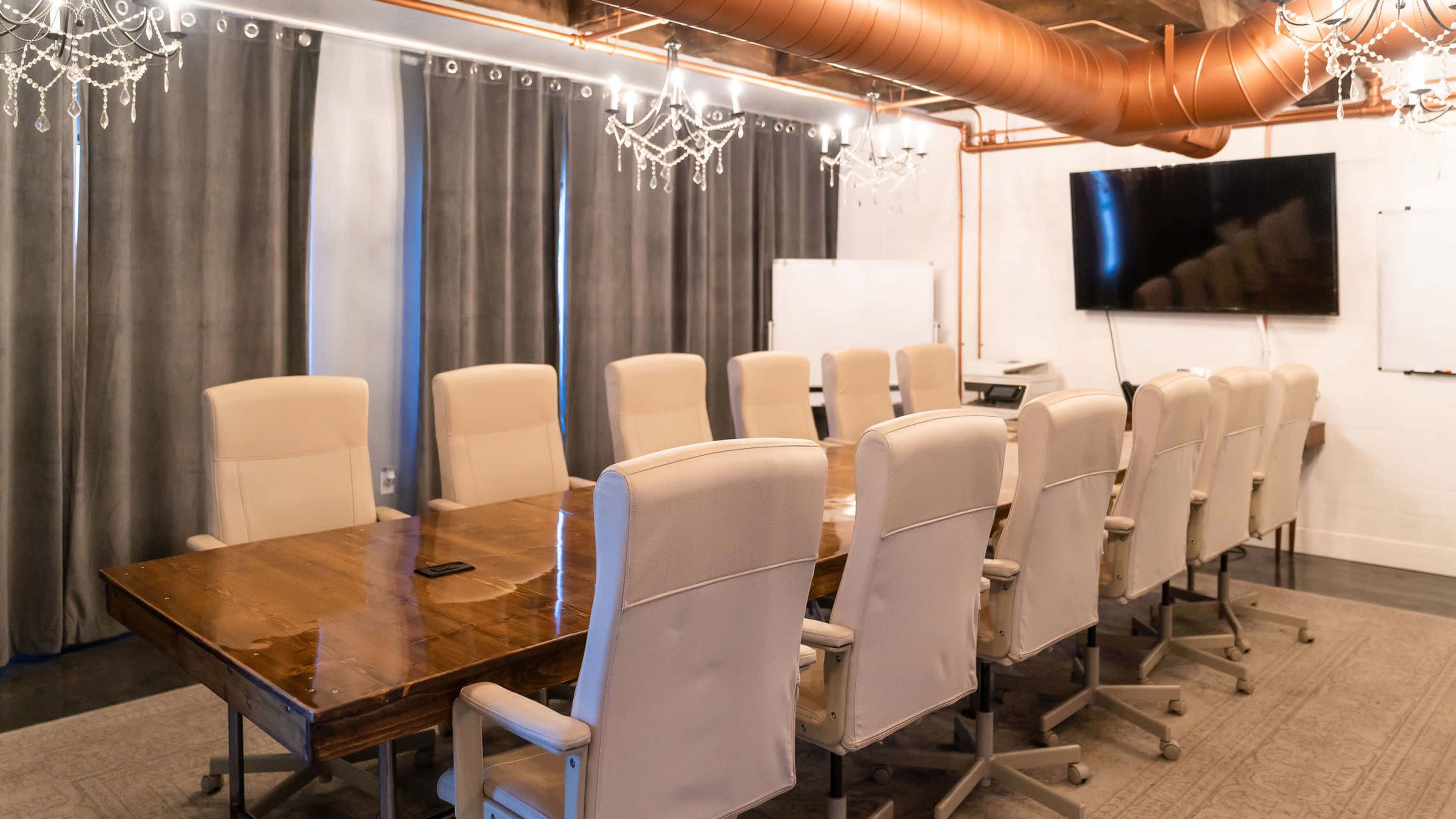 A spacious conference room features a large wooden table surrounded by twelve beige chairs, with curtains and chandeliers enhancing the decor.