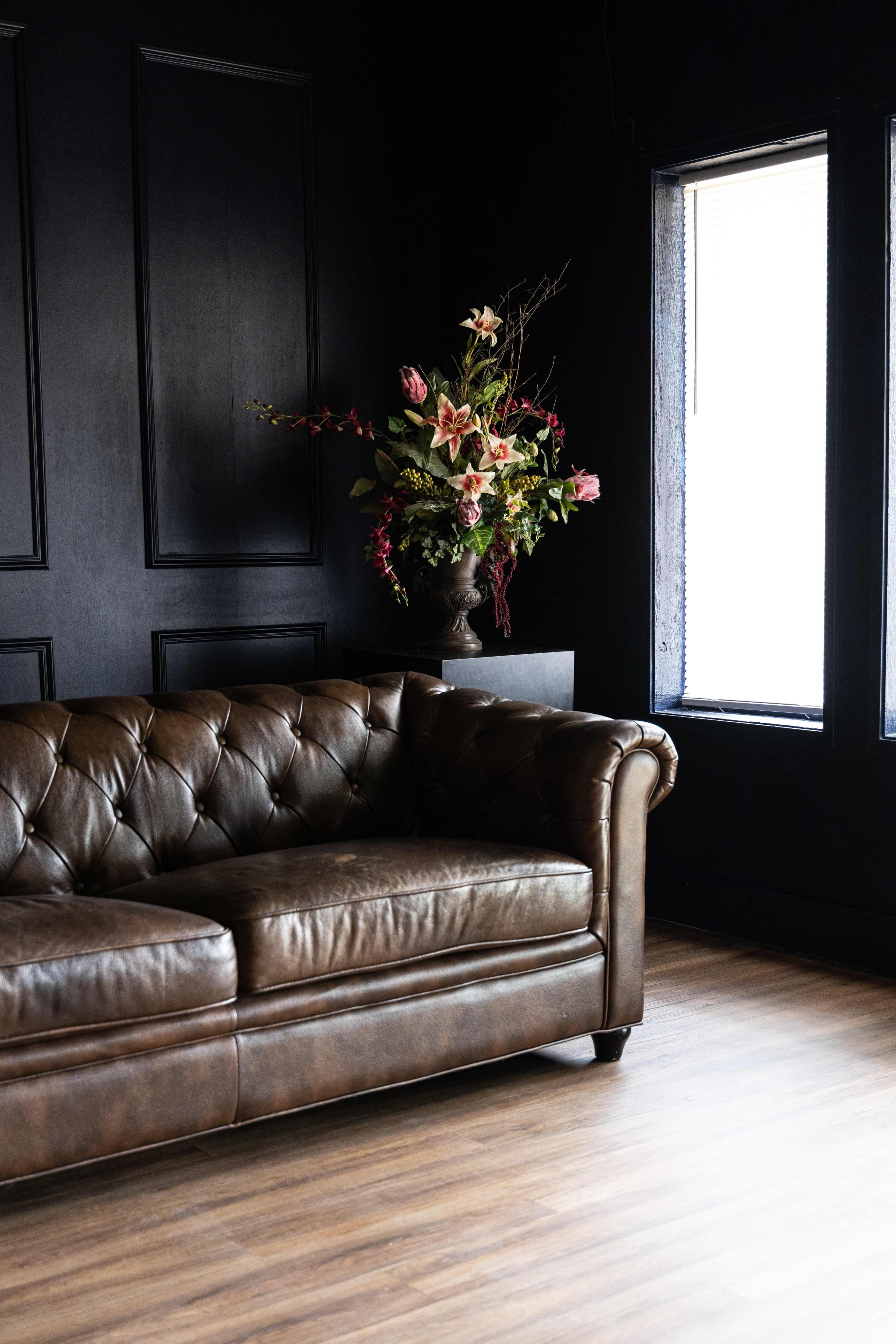 The image features a brown leather chesterfield sofa next to a decorative floral arrangement in a dark room with large windows.