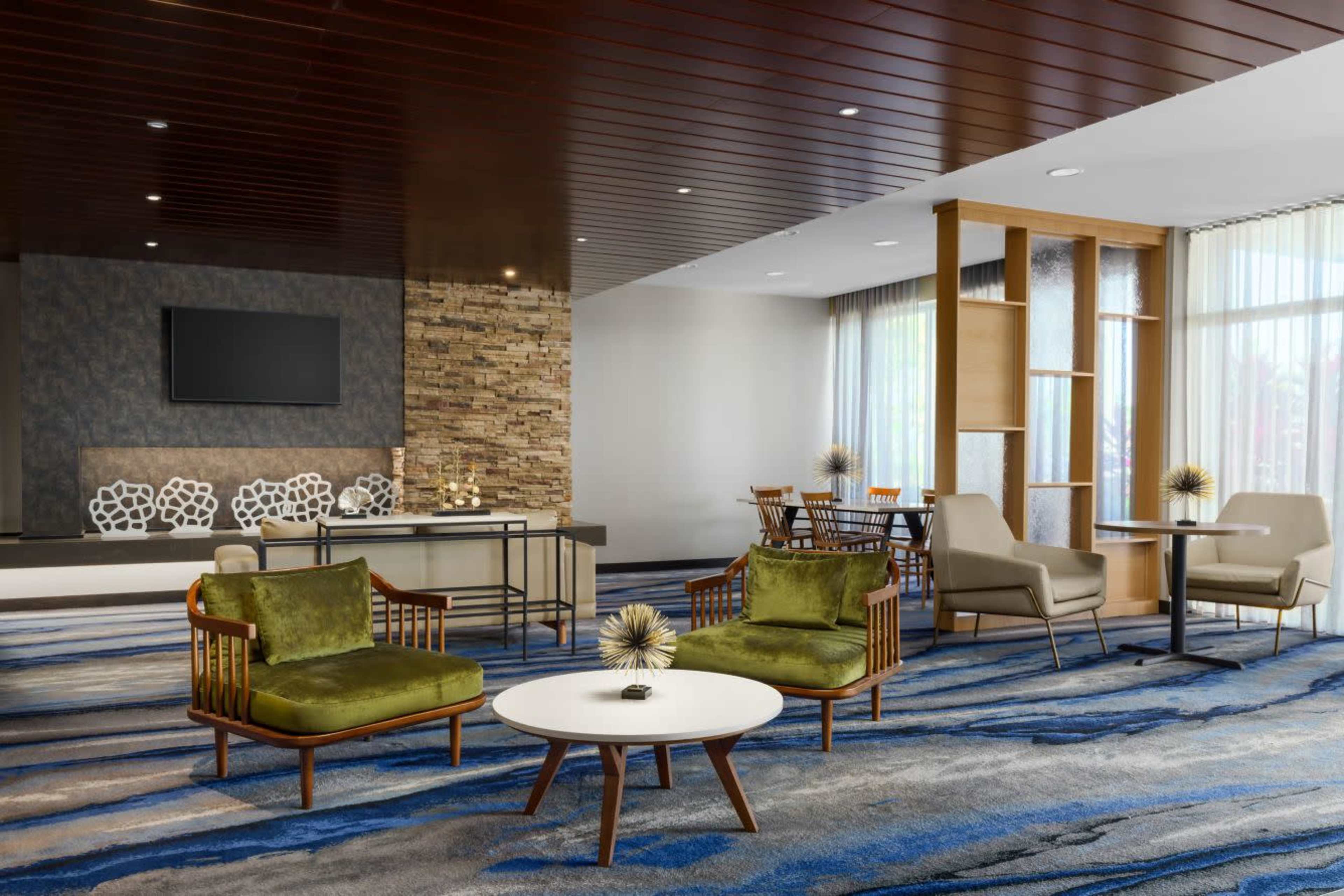 The image features a modern lobby area with seating arrangements including green armchairs and a round table, complemented by a decorative stone wall and a television.