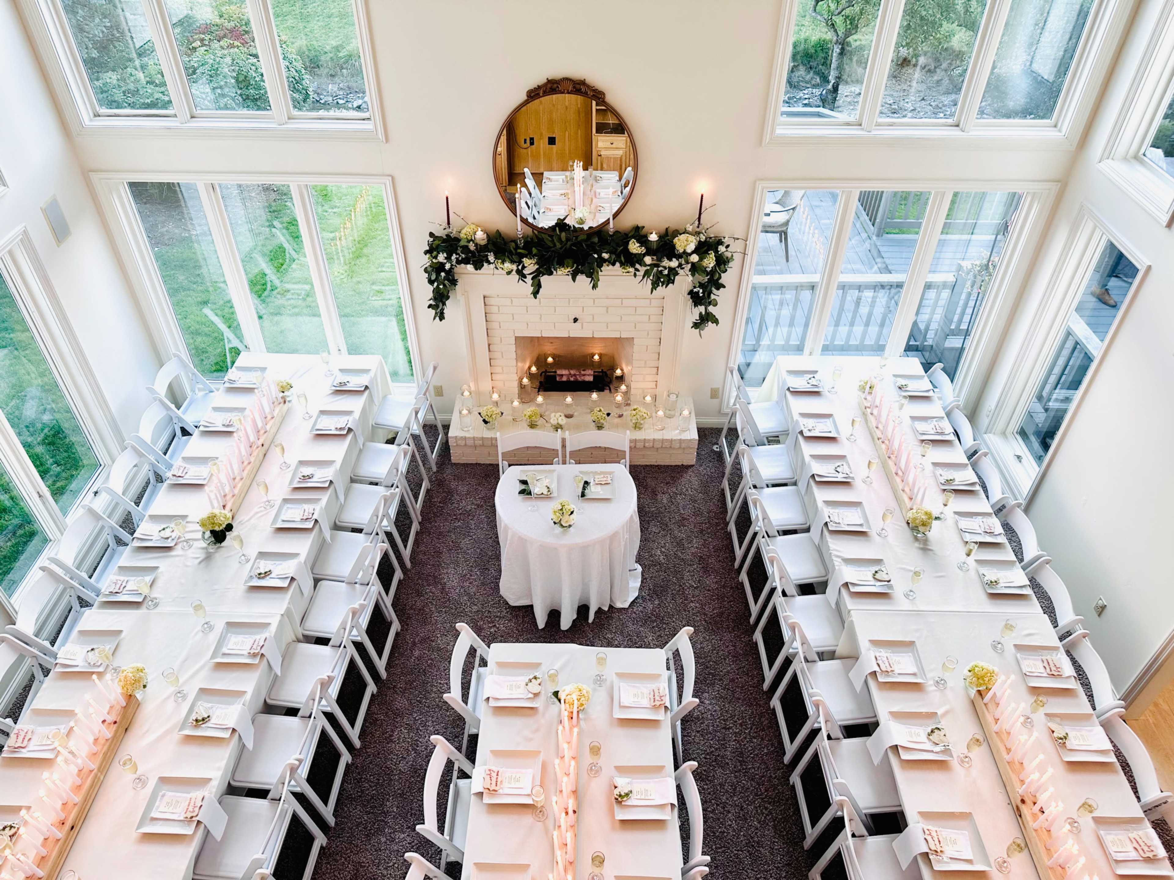 The image features a spacious dining area set with two long tables arranged in a U-shape, decorated with flowers and candles, and a fireplace adorned with greenery.