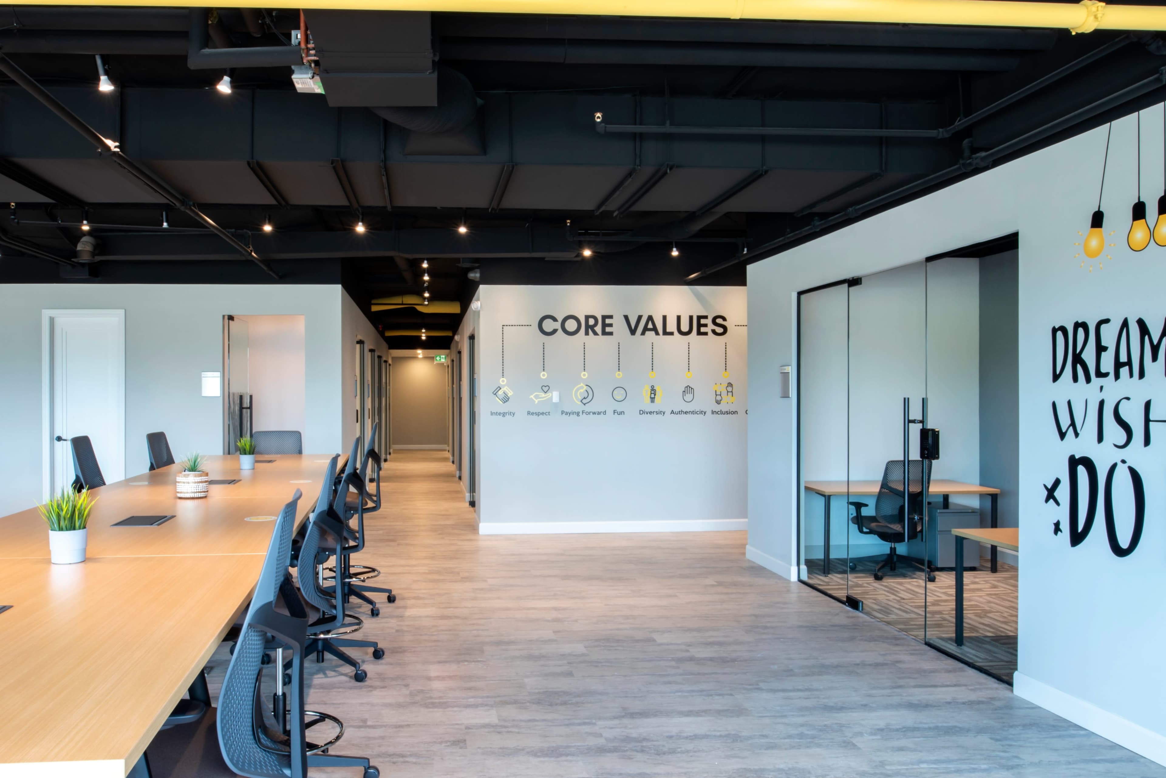 A modern office space featuring a long wooden table with black chairs, a wall displaying "CORE VALUES," and a glass-walled meeting room on the right.