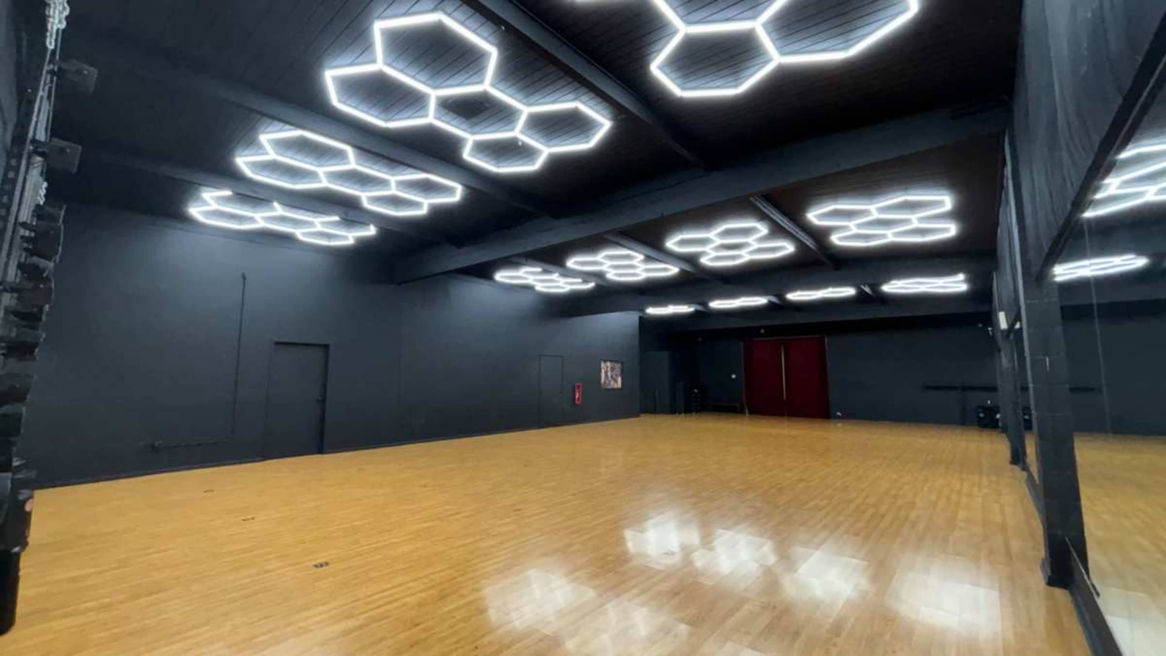 The image shows a large, empty room with hexagonal light fixtures on the ceiling and a wooden floor.