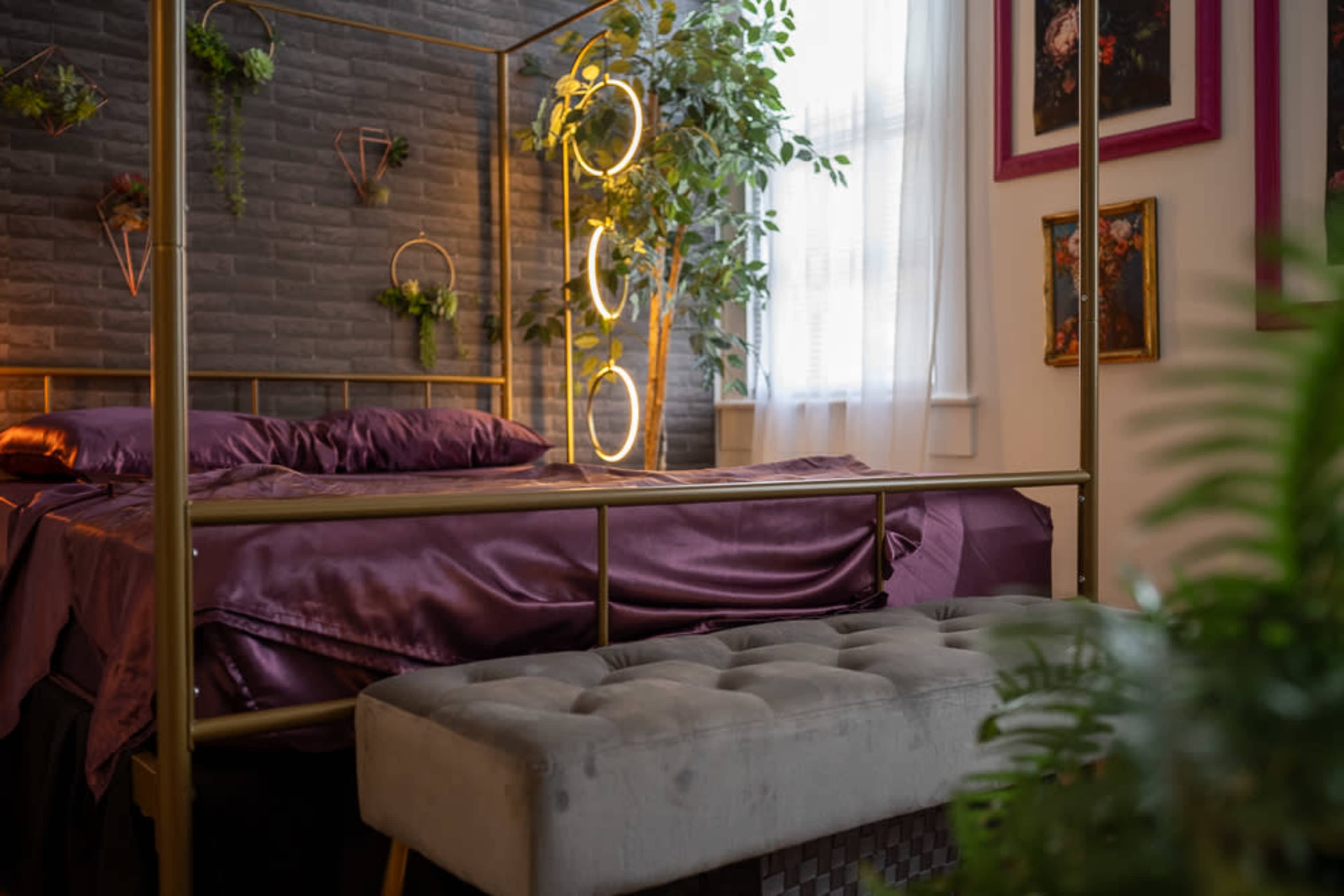 The image shows a decorative bedroom featuring a gold-framed bed with purple linens, a bench at the foot of the bed, and various plants integrated into the decor.