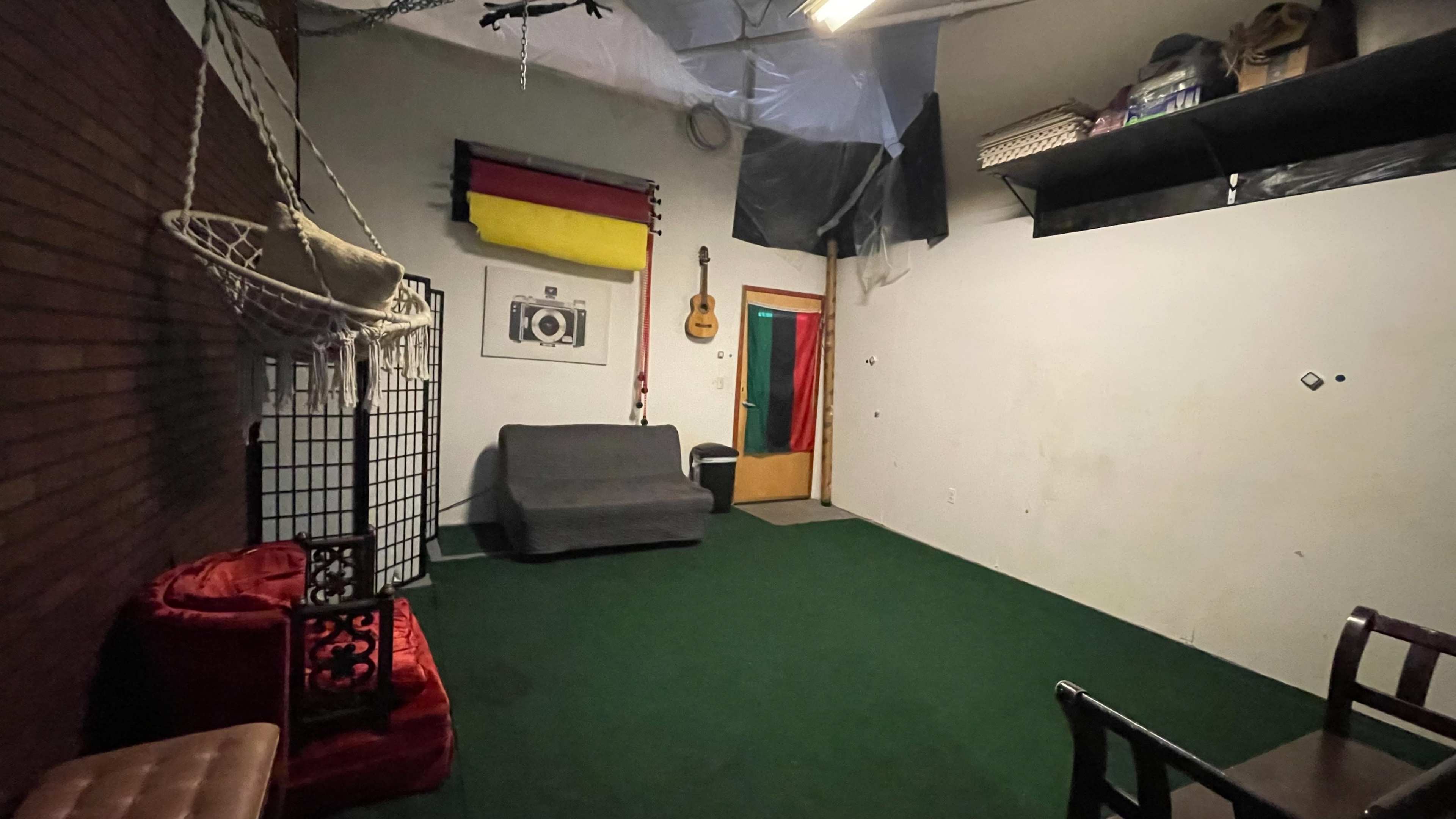 The image shows a sparsely furnished room with green carpet, a guitar hanging on the wall, and a door leading to another space.
