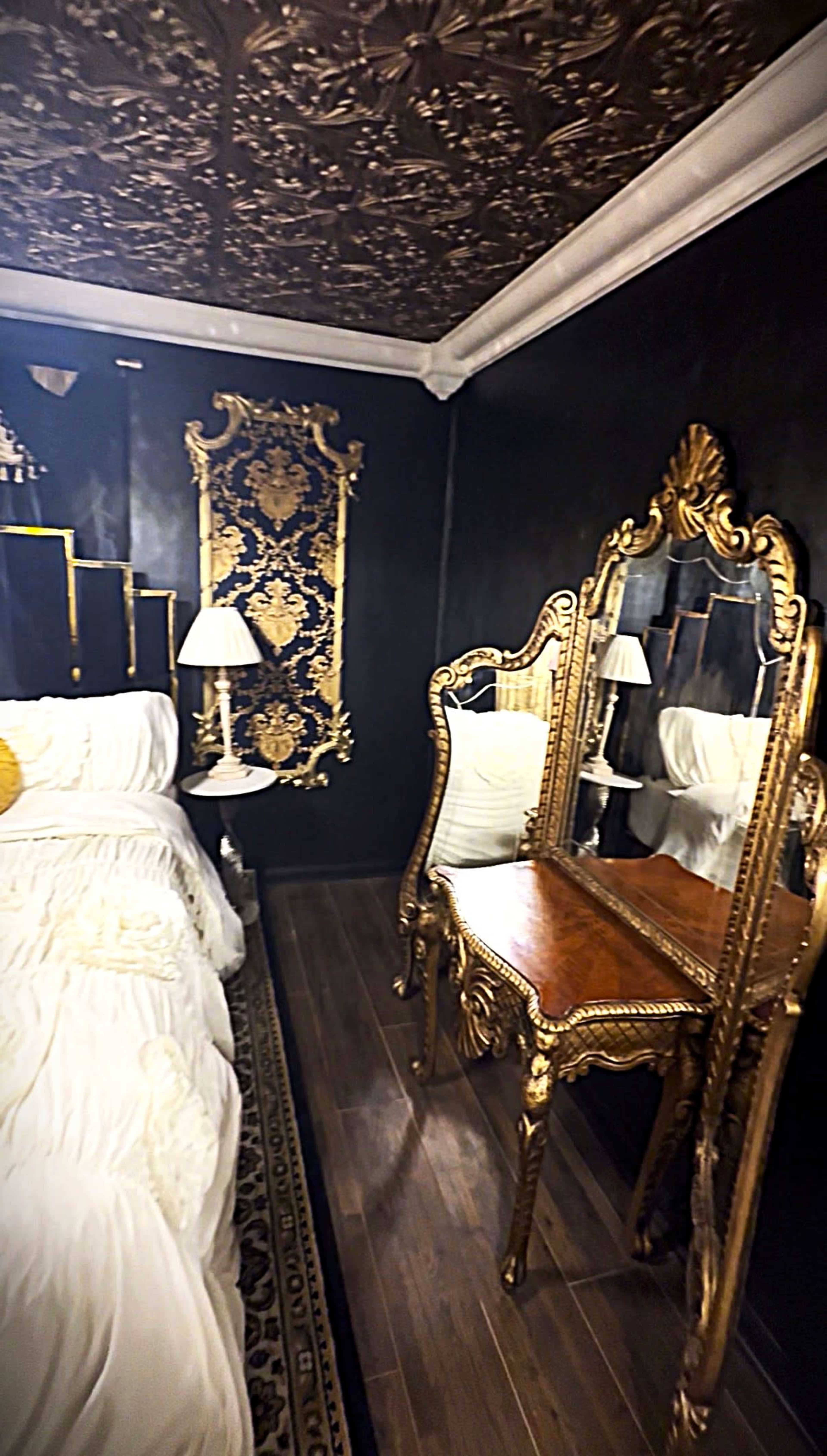 The image shows a bedroom with a dark wall, an ornate golden mirror and vanity, and a plush bed with white bedding.