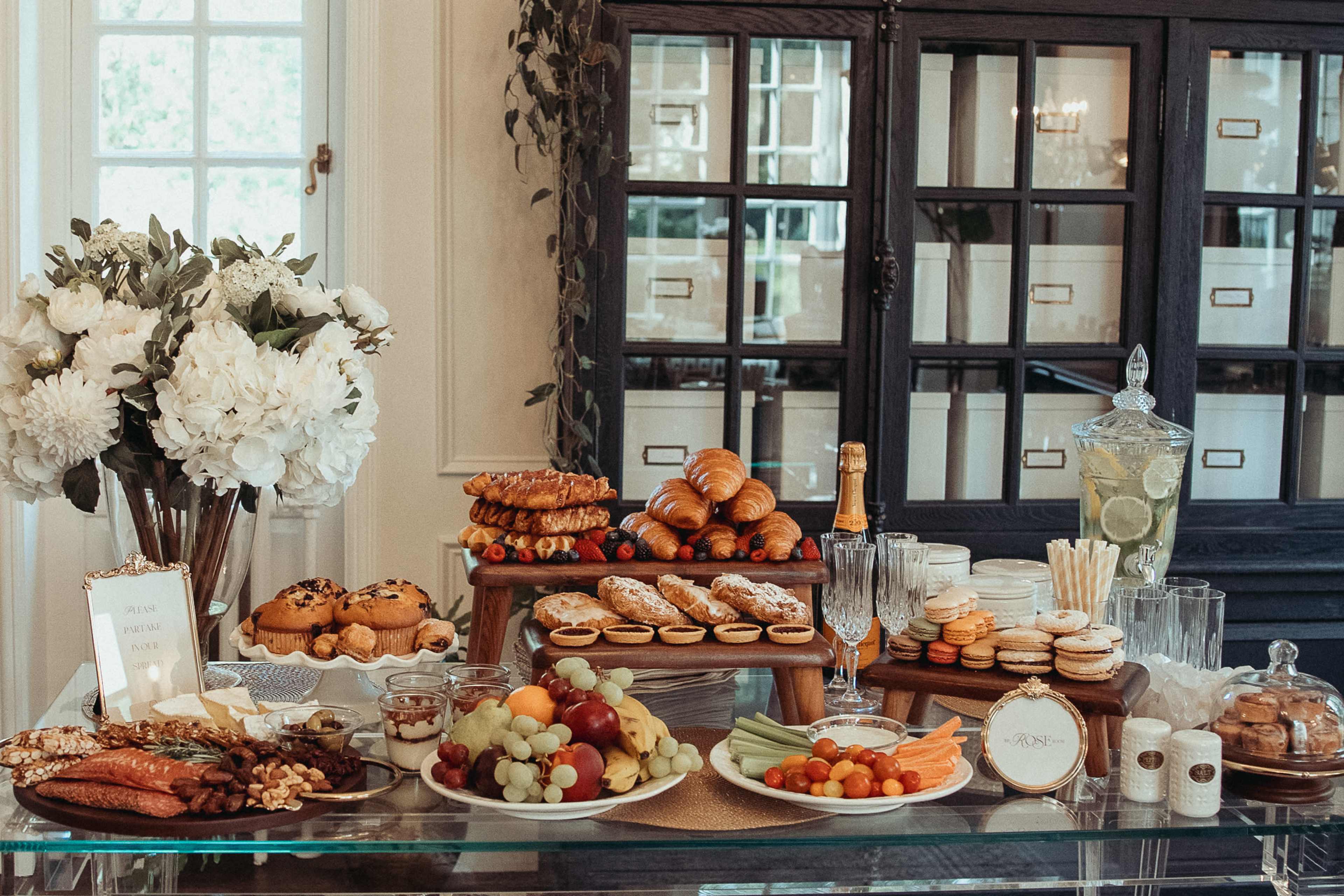 A banquet table displays an assortment of pastries, fruits, and beverages, surrounded by decorative flowers and glassware.