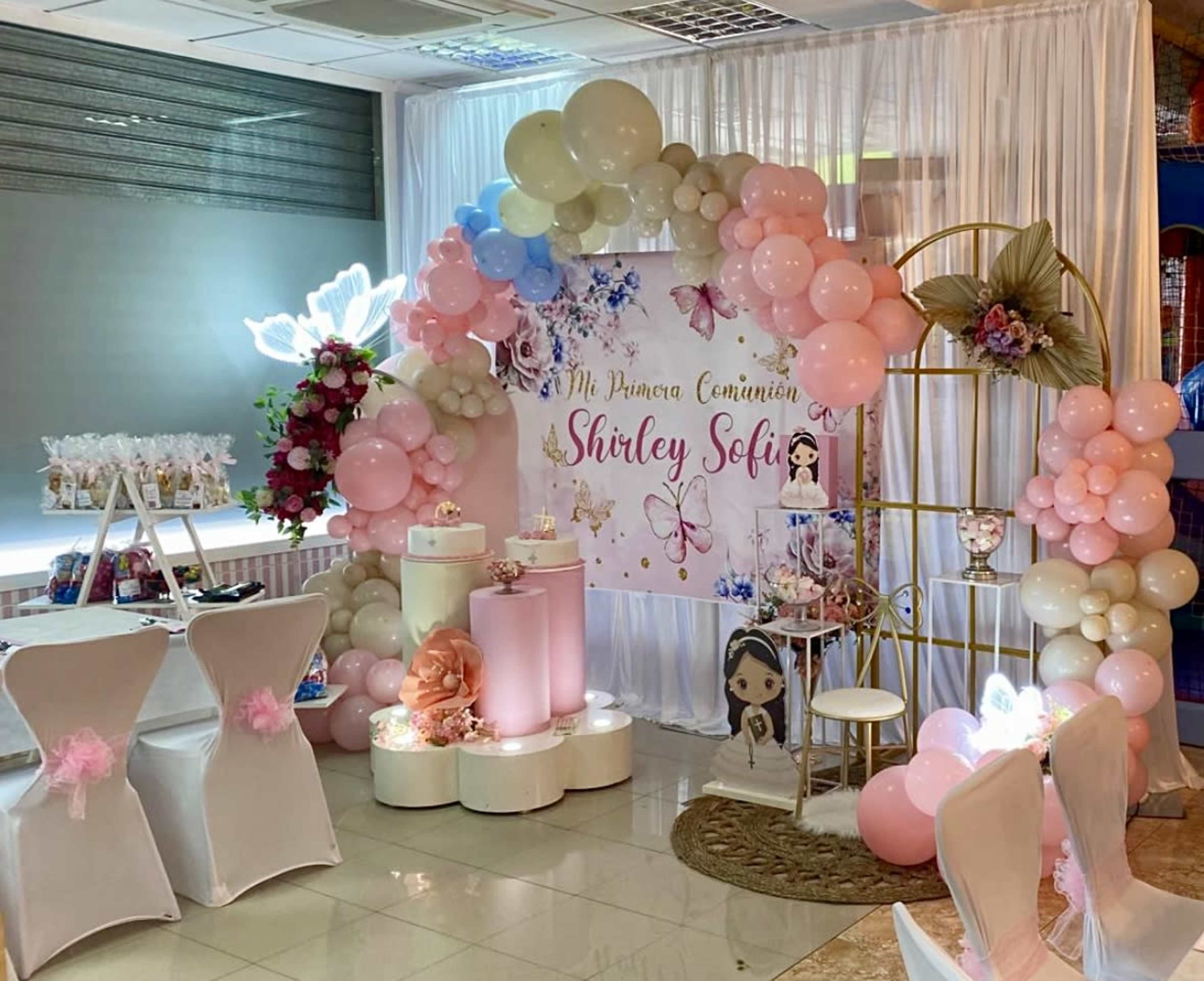 The image shows a decorated party area featuring a backdrop with balloons, a sign reading "Mi Primera Comunión Shirley Sofi," and various decorative elements such as flowers and stands.