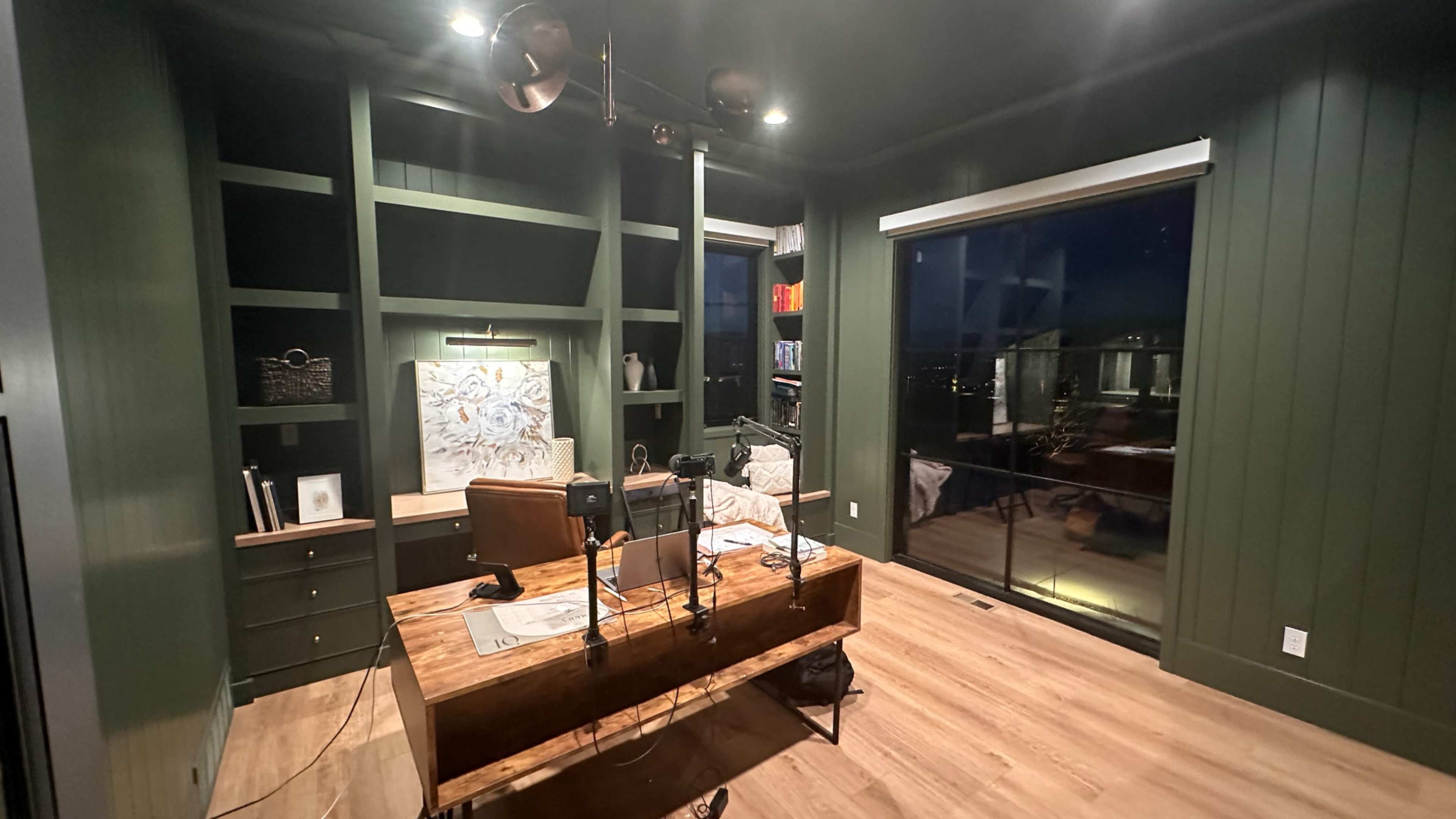 A modern home office with a large wooden desk, green walls, shelves filled with books, and a large window revealing a night view.