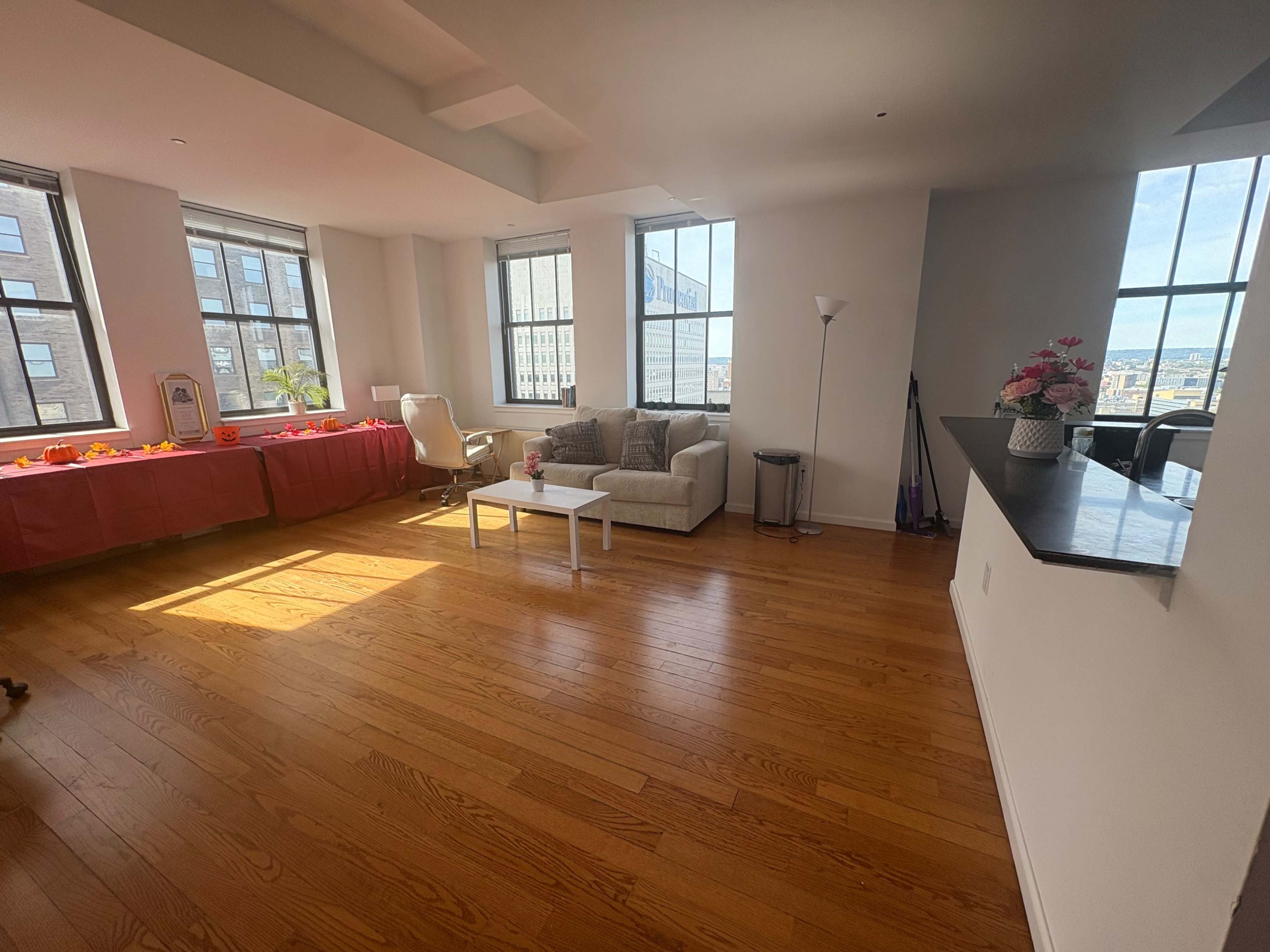 The image shows a spacious, well-lit living area with a couch, a small white coffee table, and large windows, complemented by wooden flooring and minimal furnishings.