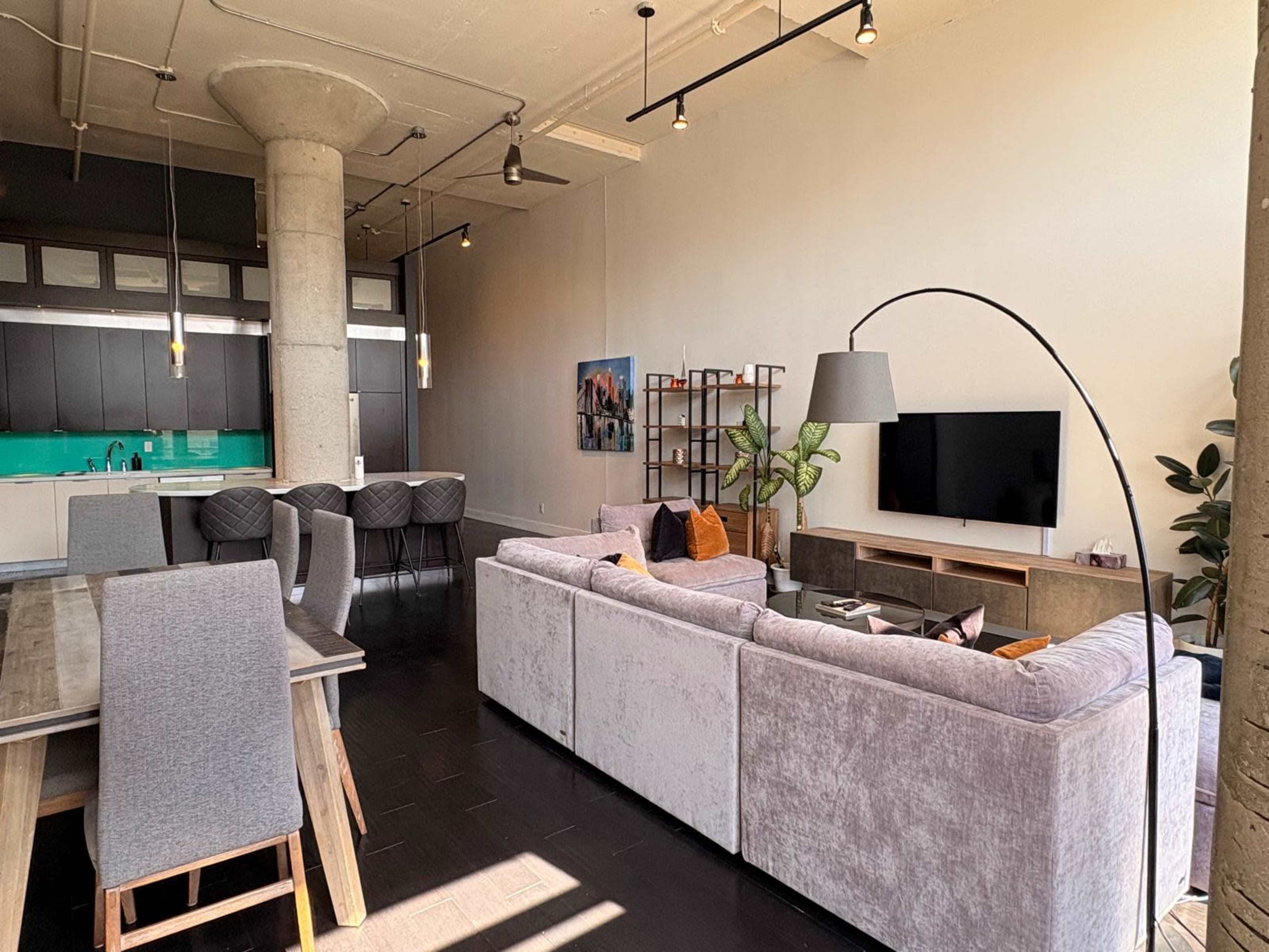 The image shows a modern living room featuring a large gray sofa, a television on a wooden stand, and a dining table with chairs, all set in an open-concept space with a kitchen in the background.