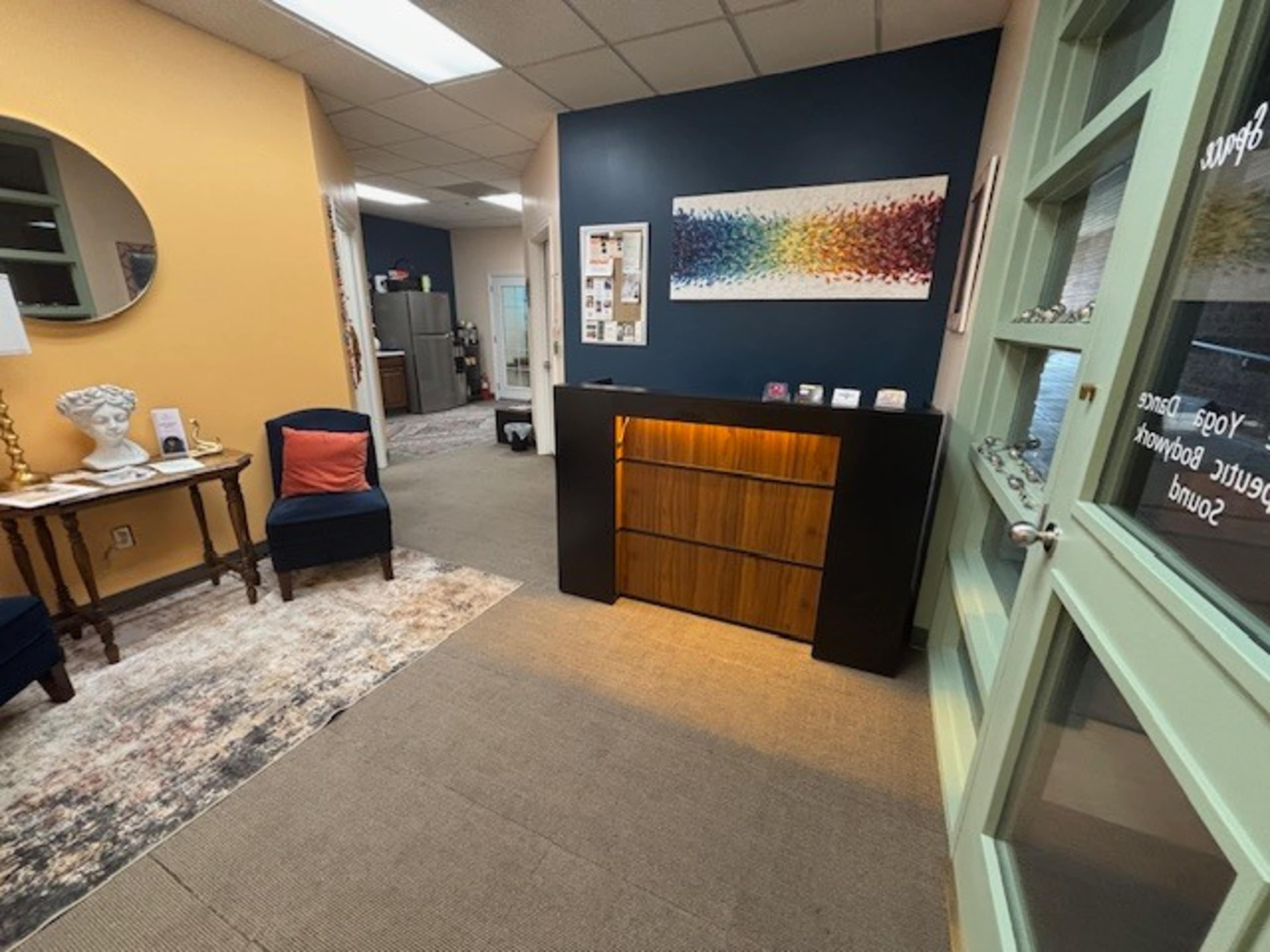 The image shows a welcoming interior space with a decorative mirror, a wooden bookshelf with lighting, and chairs, along with a wall featuring colorful artwork.