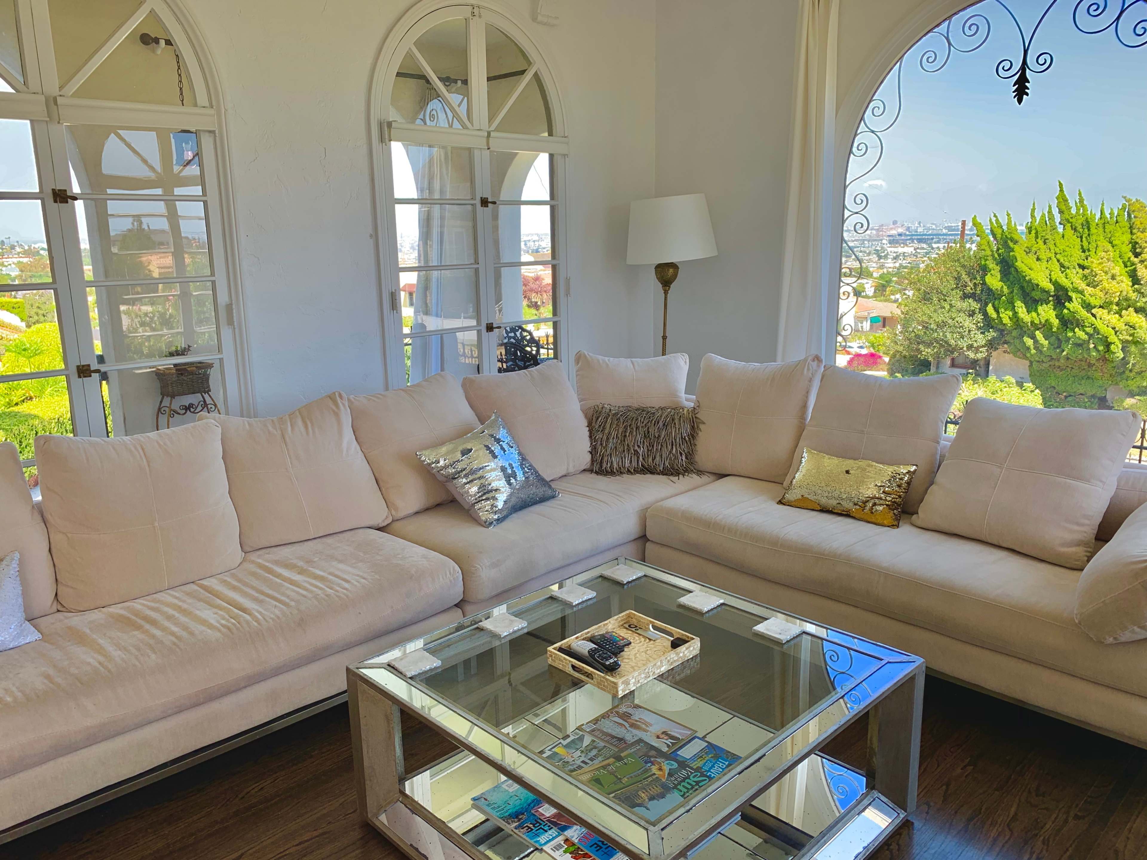 The scene features a light-colored sectional sofa with decorative pillows and a glass coffee table in a sunlit living room overlooking a garden through arched windows.