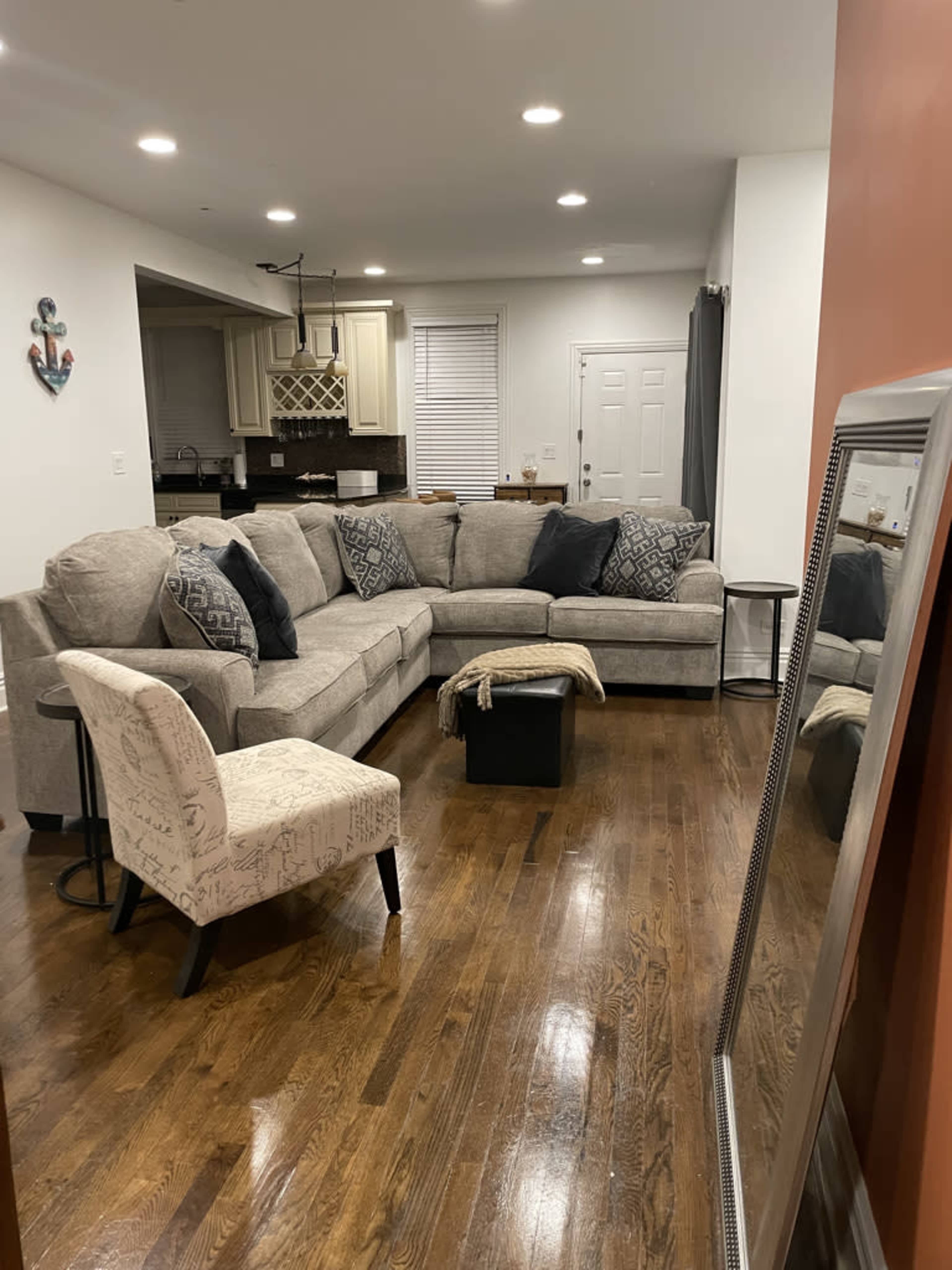 A cozy living room features a large sectional couch, an accent chair, a coffee table, and a mirror, with hardwood flooring and a view into a kitchen area.