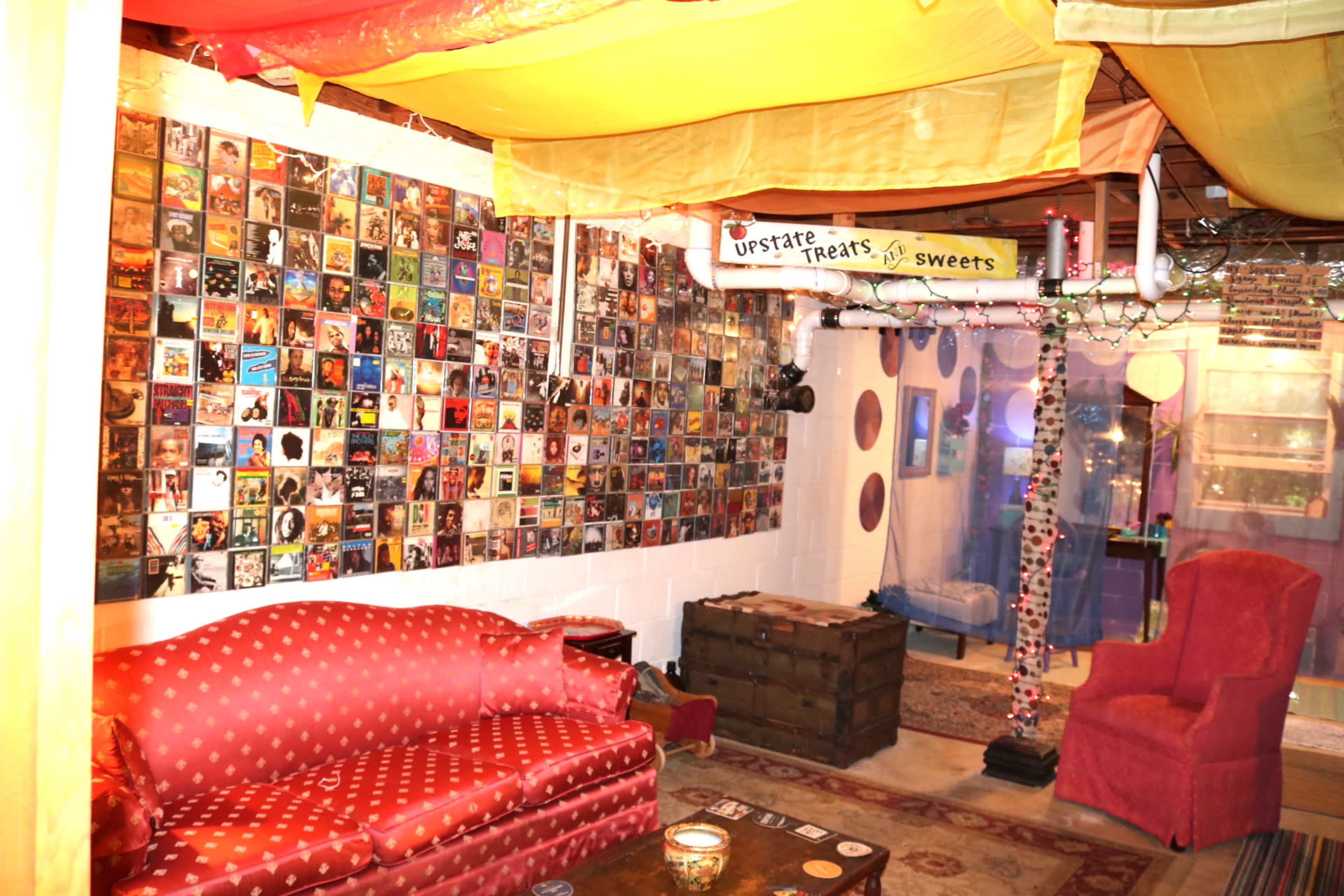 A cozy room decorated with a wall covered in CDs, a red couch, a pink chair, and colorful fabric draping from the ceiling.