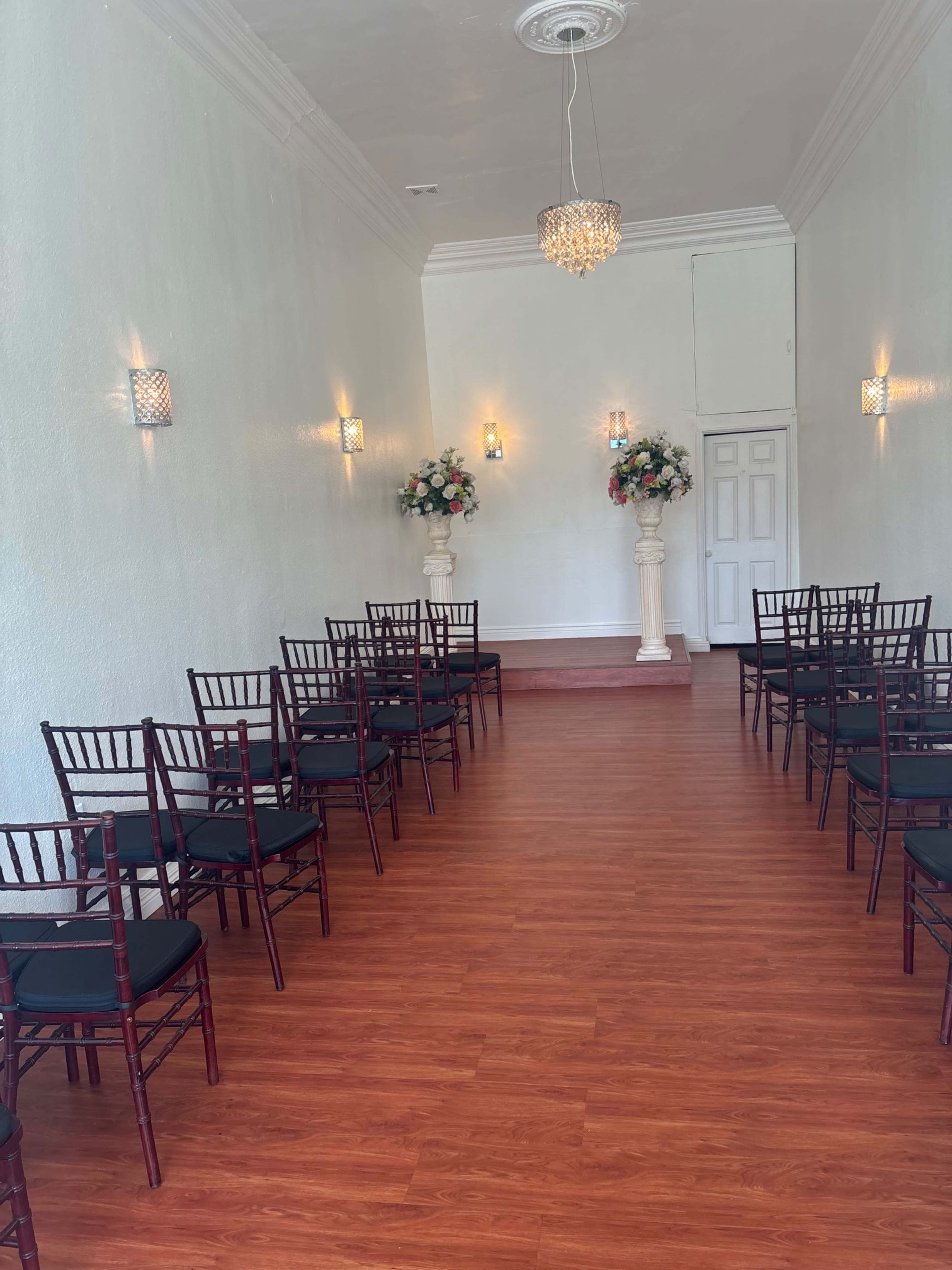 The image shows a bright, semi-furnished room with rows of dark chairs facing a decorative floral arrangement on pedestals at one end.