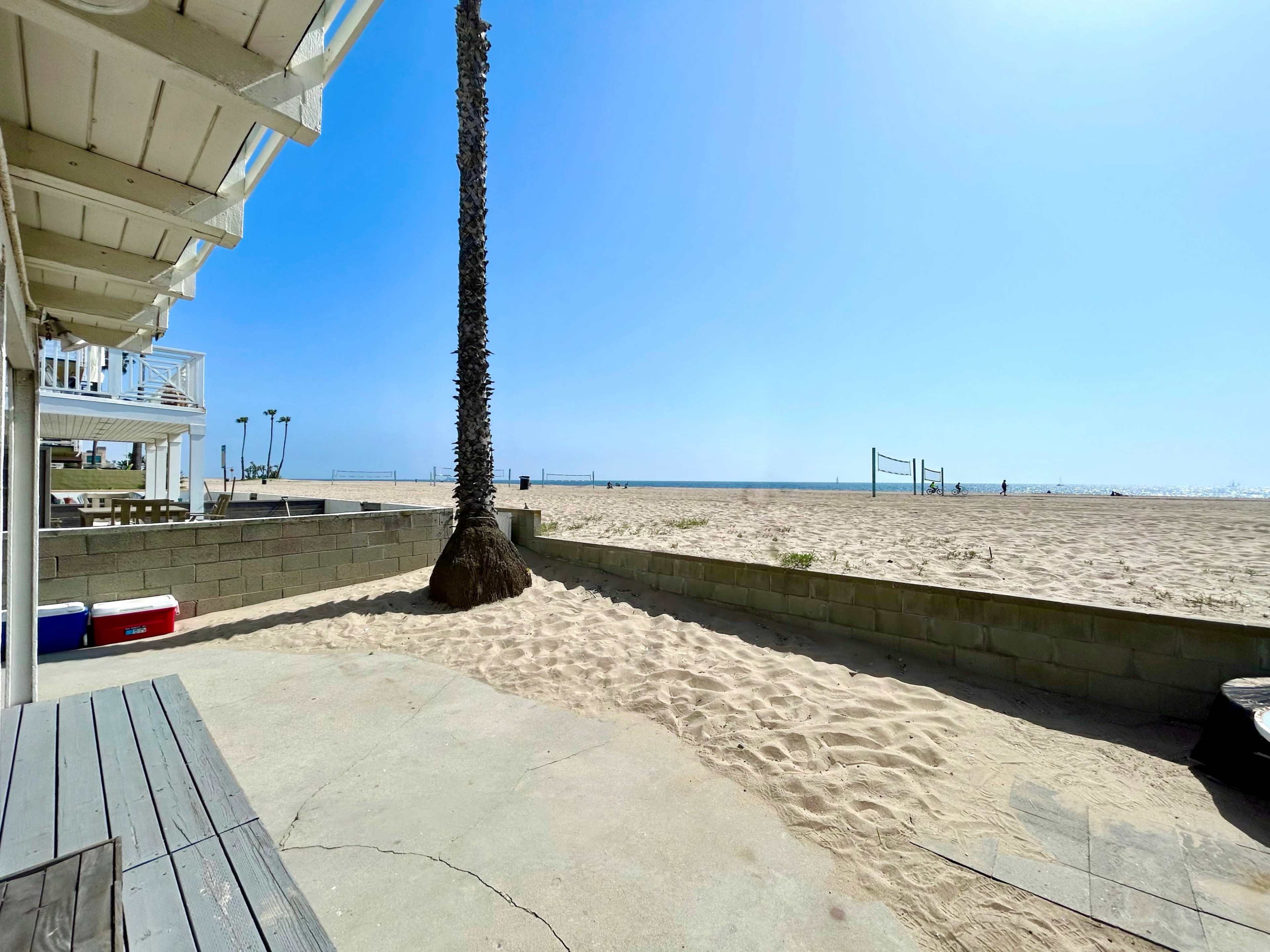 A sandy beach with volleyball nets in the distance, viewed from a patio next to a palm tree.