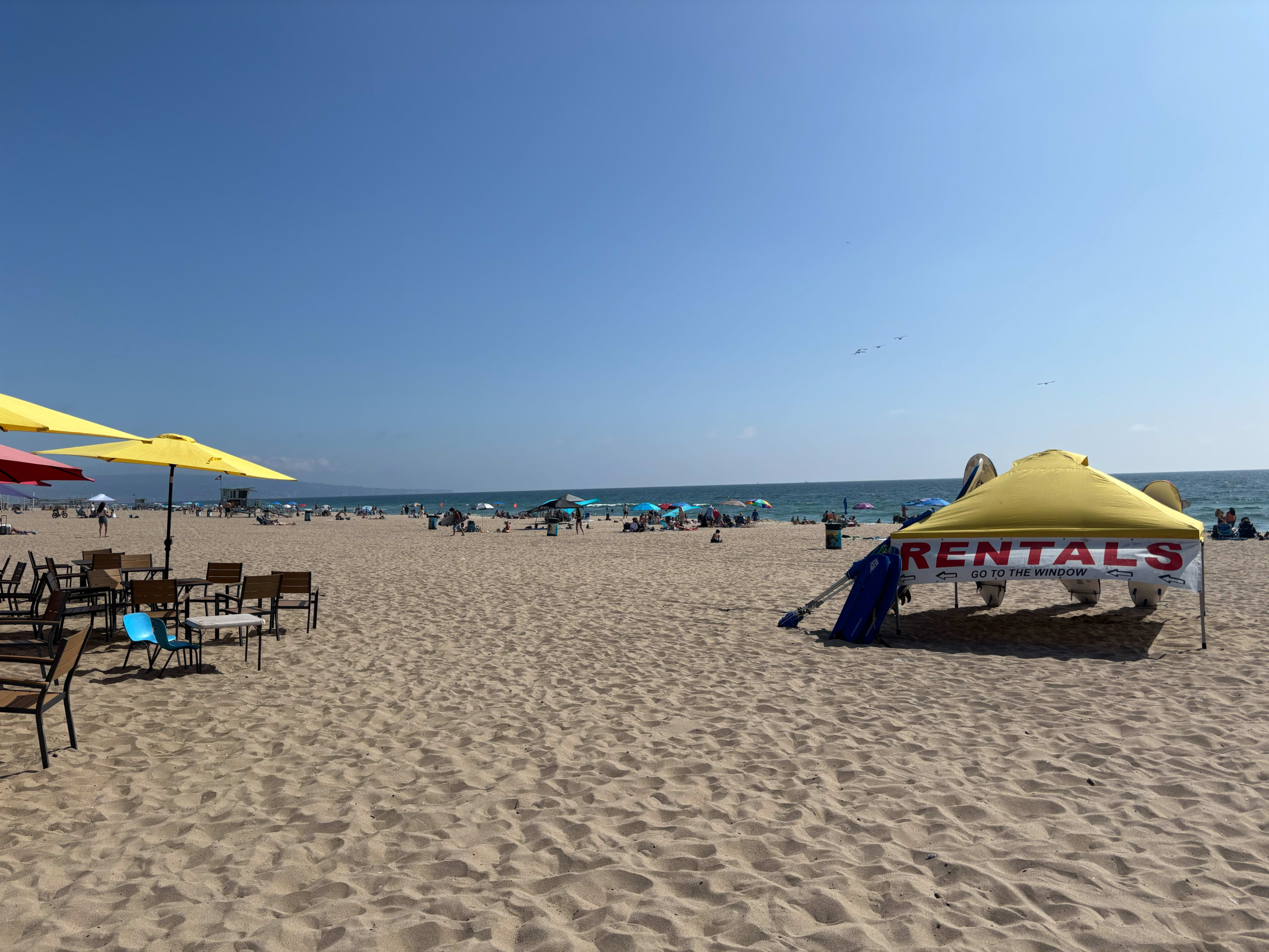 The image shows a busy beach with umbrellas, people sunbathing, and a rental tent set up on the sand.