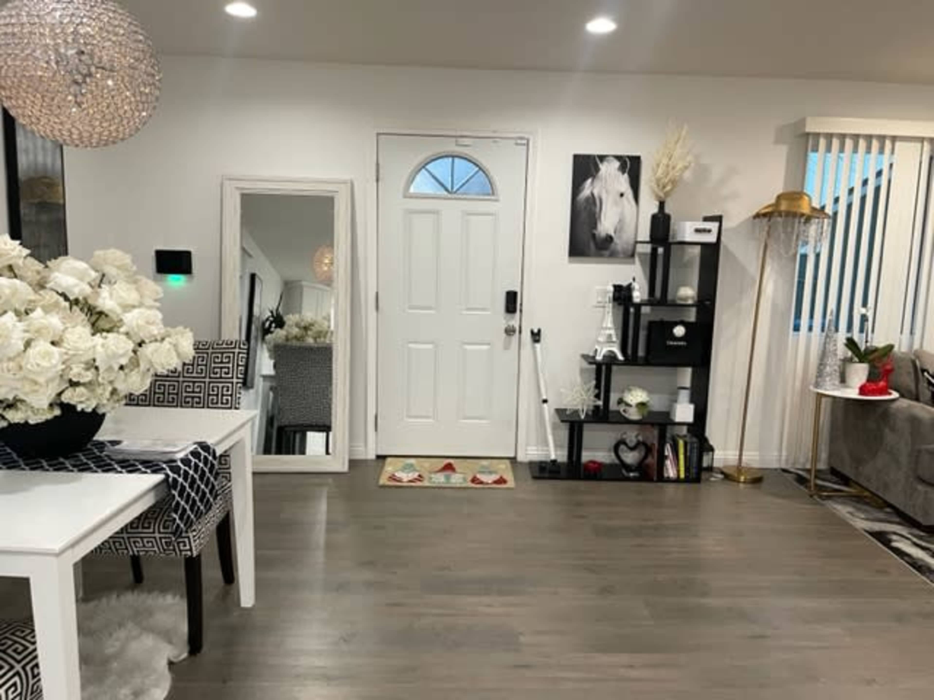 A modern living space with a white front door, a mirror, a dining table with flower arrangements, and a decorative shelf against the wall.