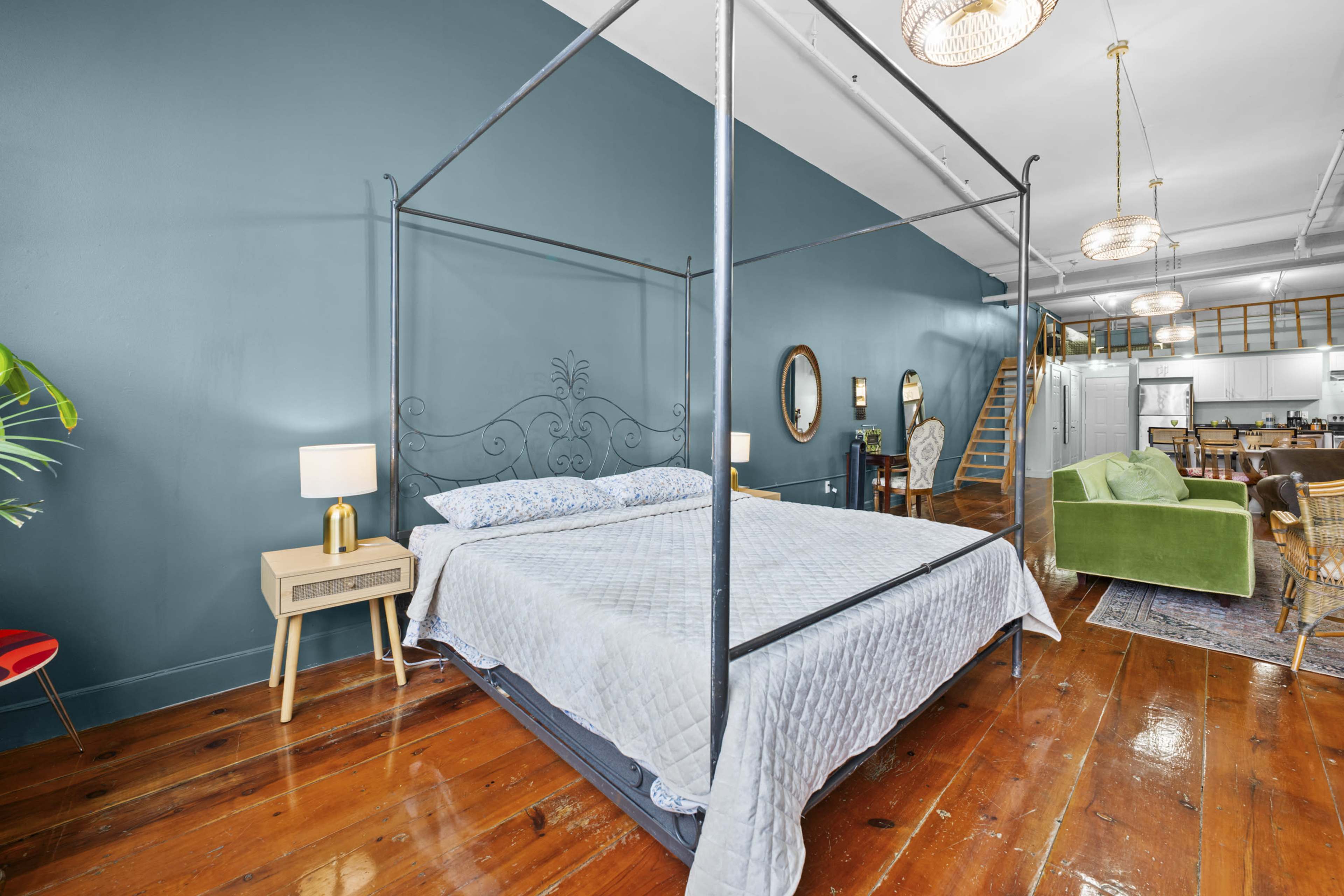 The image shows a spacious bedroom featuring a metal canopy bed, wooden floors, and a teal accent wall, with additional living space visible in the background.