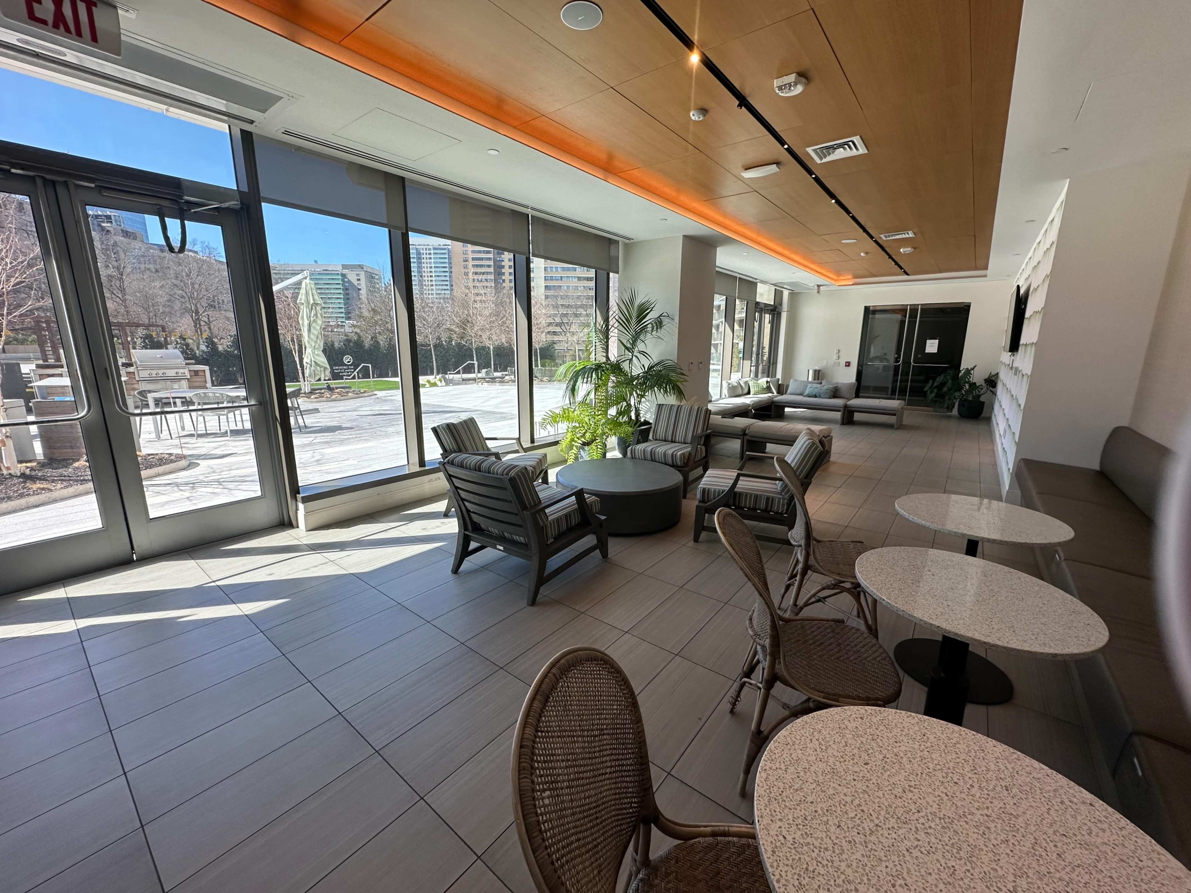 The image shows a modern lounge area with comfortable seating, large windows, and a view of an outdoor patio space.