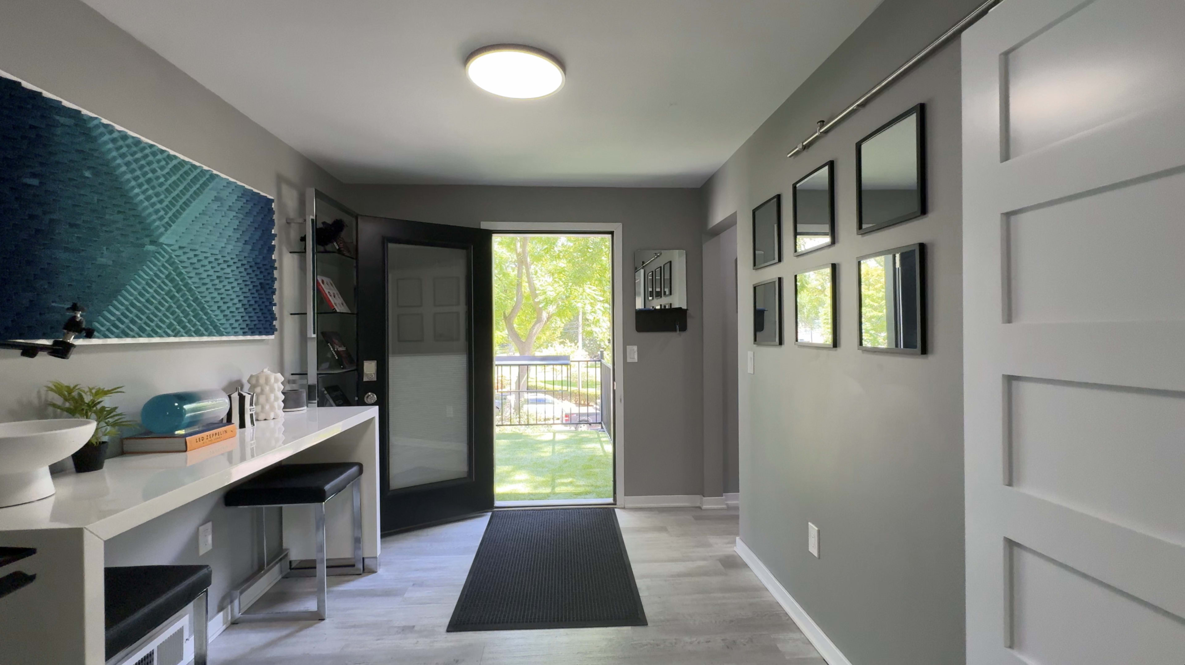 The image shows a modern entryway with a black front door open to a view of greenery, complemented by a white countertop and black stools along one wall, and several framed pictures on the opposite wall.