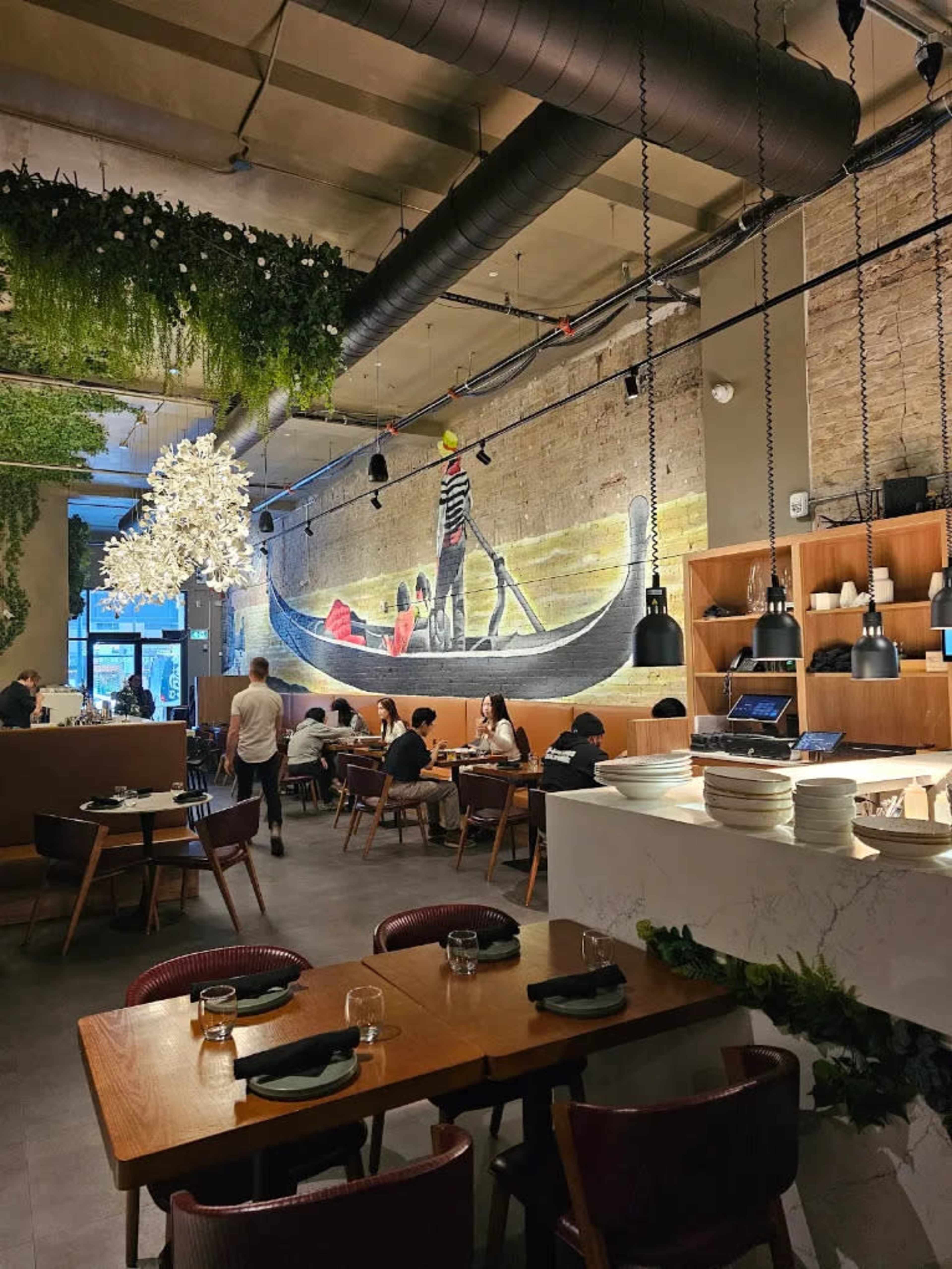 The image shows a modern restaurant interior featuring a mural of a gondola on the wall, with patrons seated at wooden tables under pendant lighting.