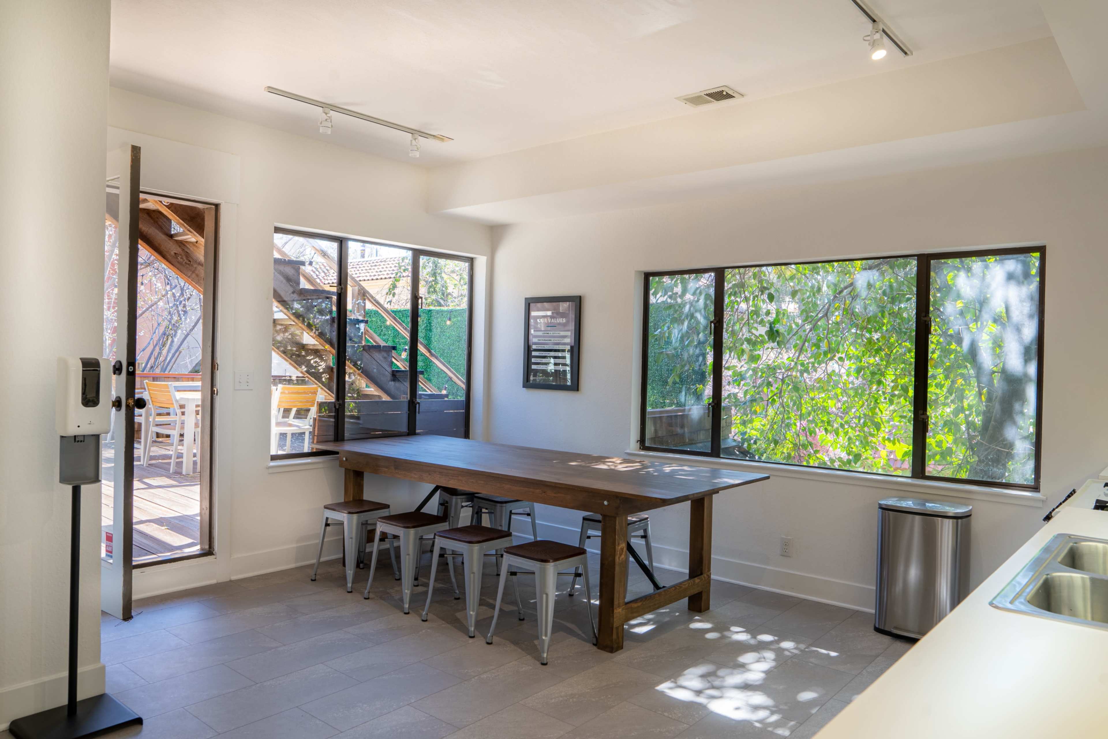 A modern dining area with a wooden table and metal stools, illuminated by natural light from large windows.