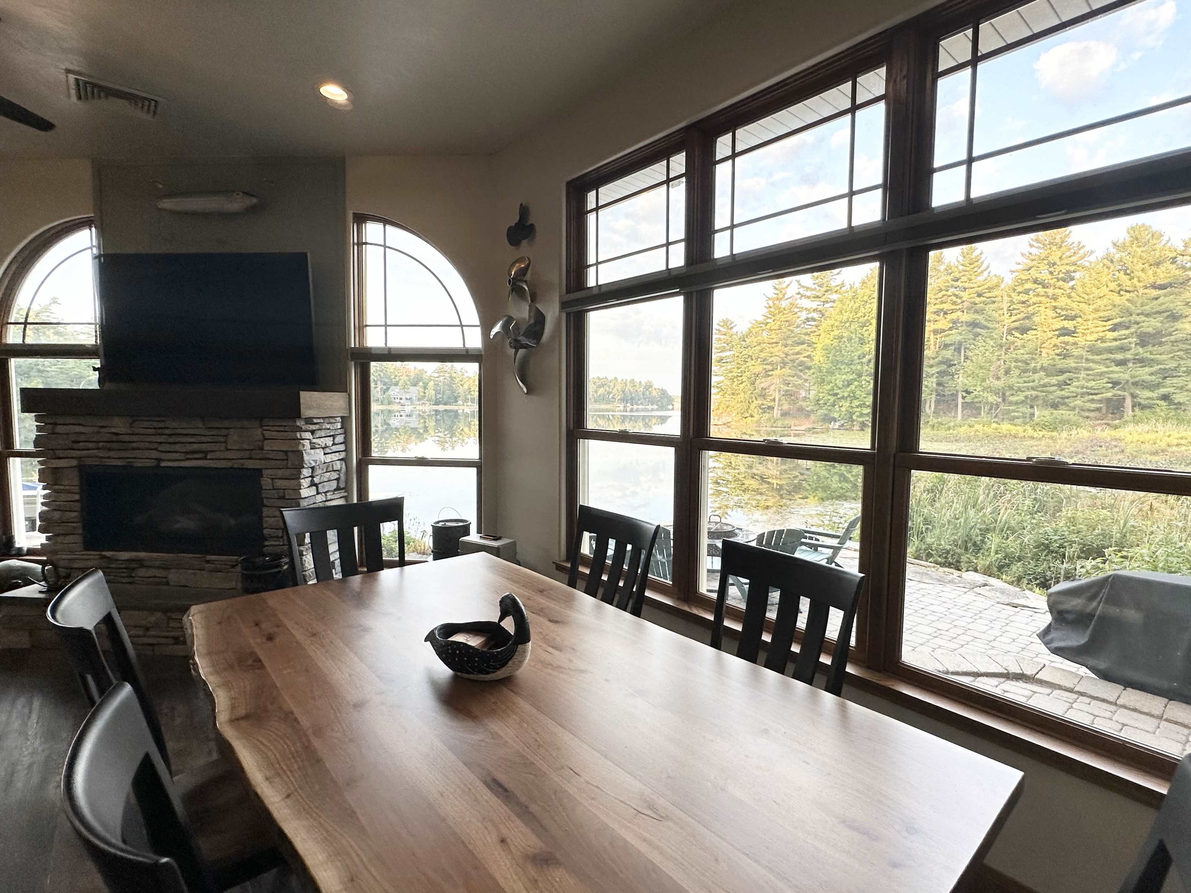 A wooden dining table is set beneath large windows overlooking a scenic view of water and trees.