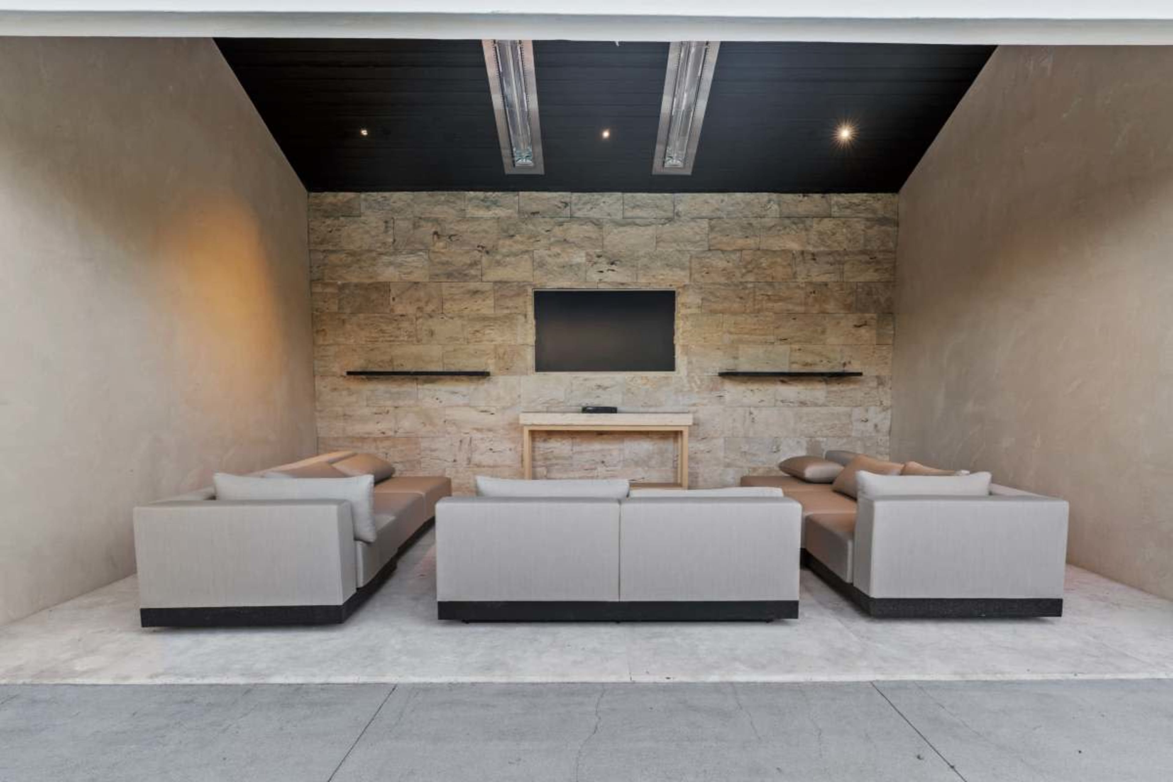 The scene features a modern outdoor lounge area with a stone wall, a large TV mounted above a wooden table, and sectional sofas arranged around it.