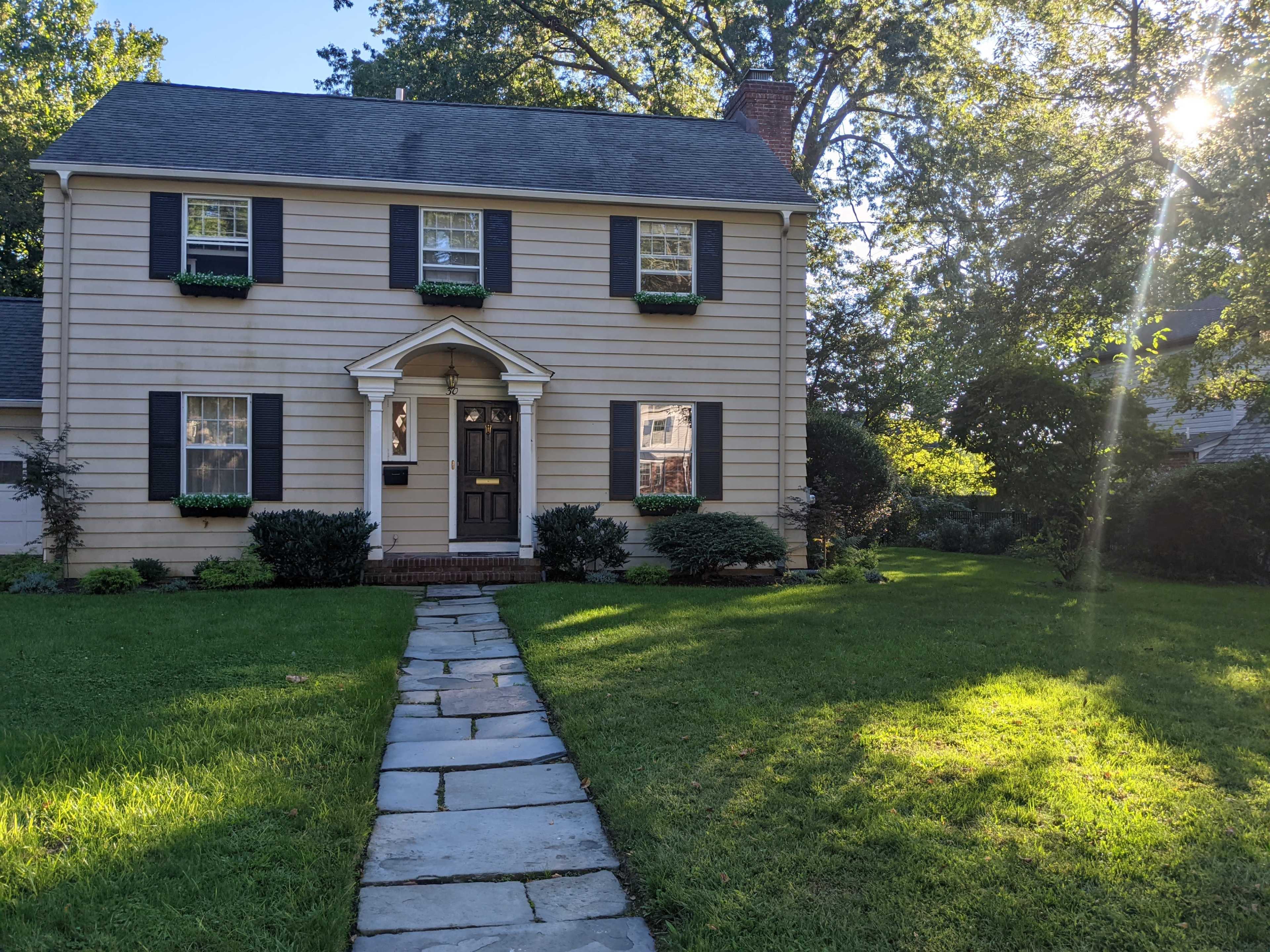 A two-story house with gray siding, black shutters, and a front walkway lined with stone leads to the entrance.