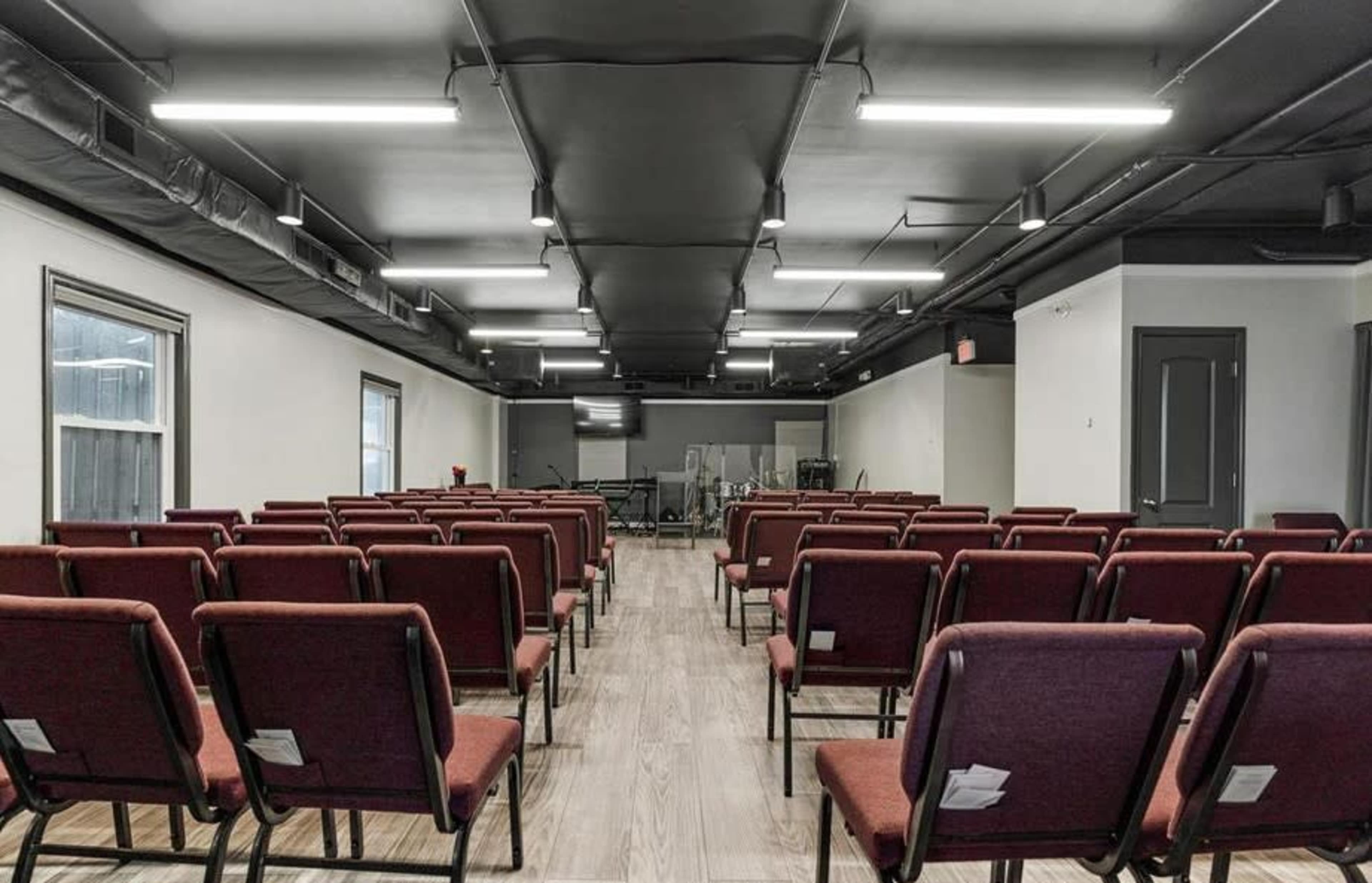 An interior space arranged with red chairs facing a stage area, illuminated by overhead lighting.